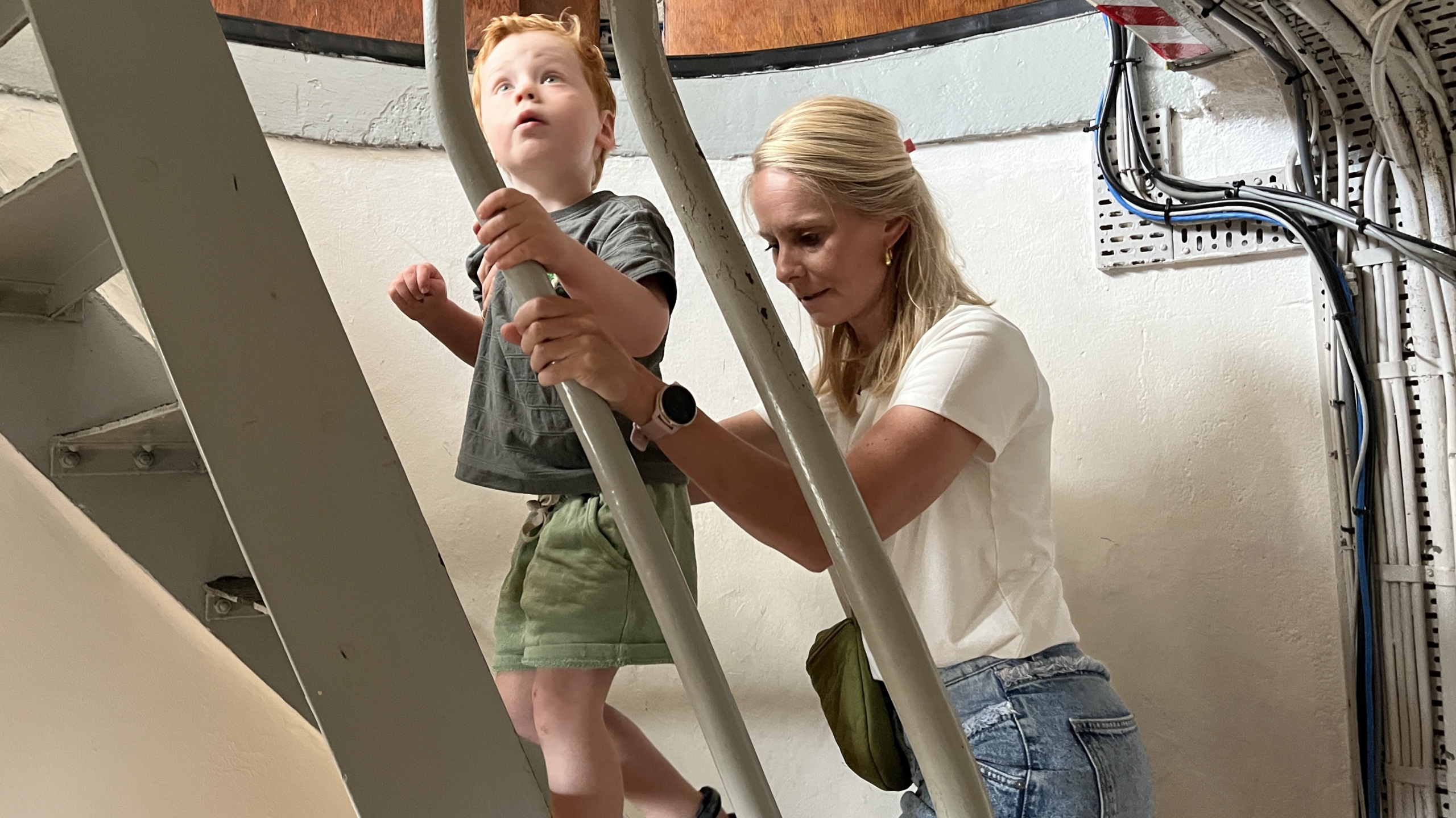 Een vrouw helpt een jong kind een steile metalen trap op te klimmen in een gebouw met blootliggende kabels langs de muur.