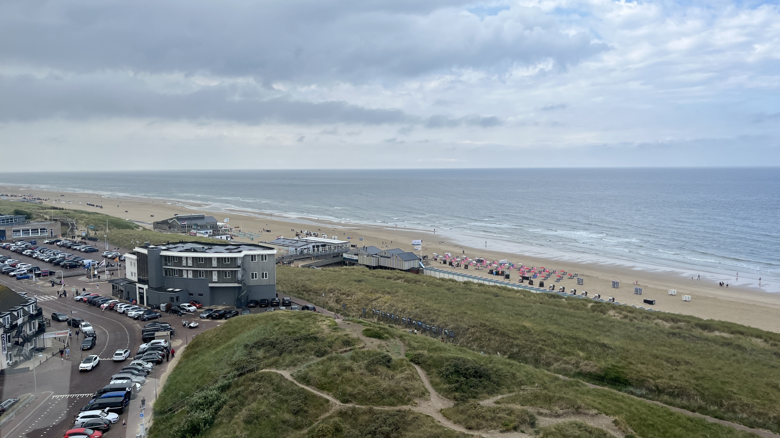 Zicht op een kustlijn met een zandstrand, ligstoelen en parasols. Er zijn enkele strandpaviljoens en parkeergelegenheden te zien. De zee strekt zich uit tot aan de horizon onder een bewolkte lucht.