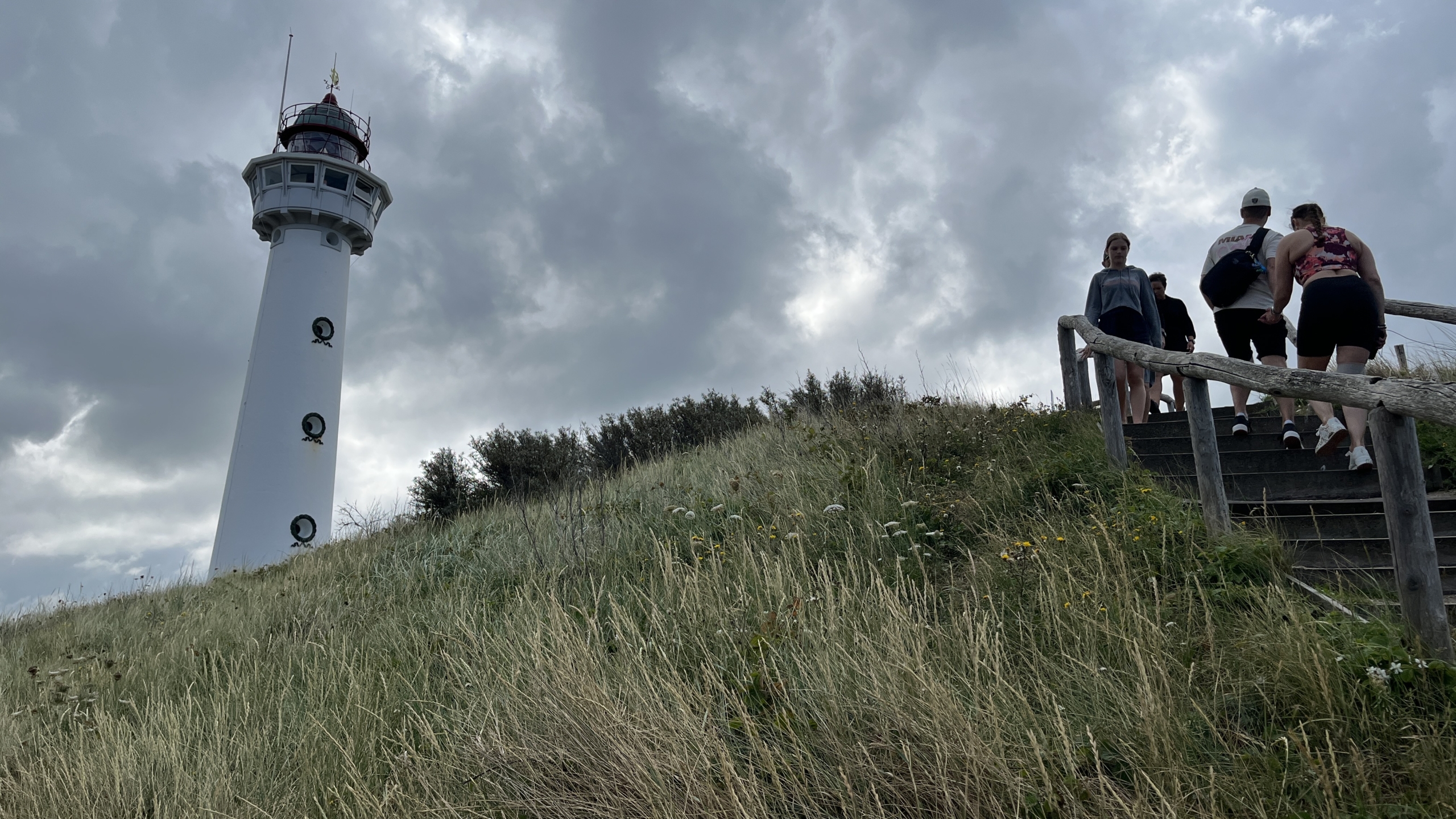 Lighthouse on a cloudy day beside a grassy hill with a group of people descending wooden stairs.