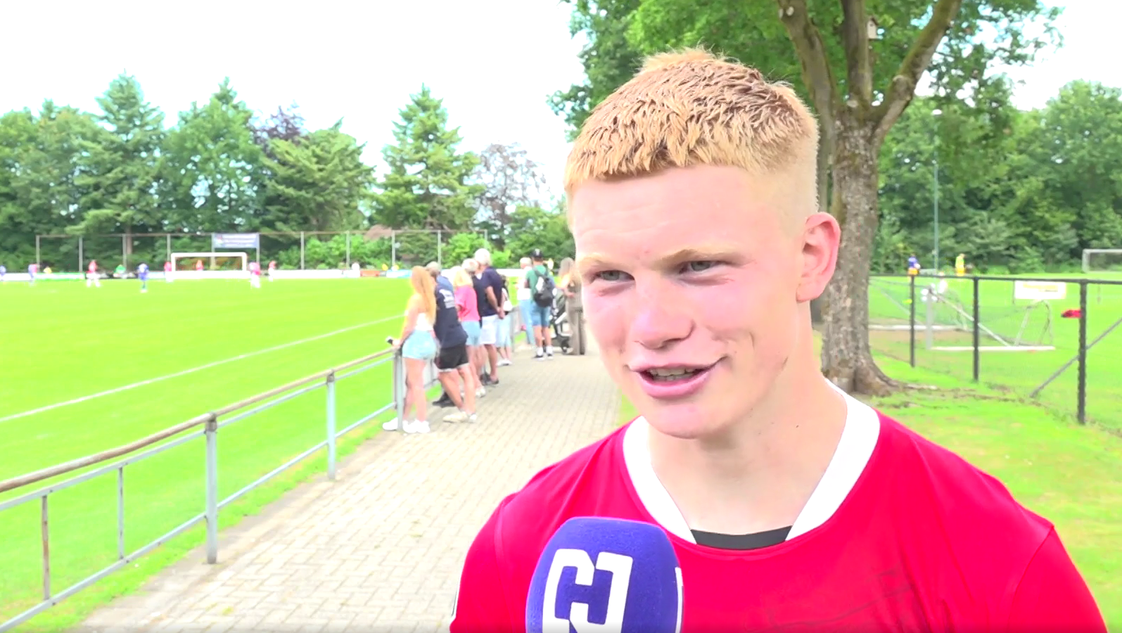 Een voetballer in een rood shirt geeft een interview met een microfoon voor een groen grasveld, omringd door toeschouwers en bomen.