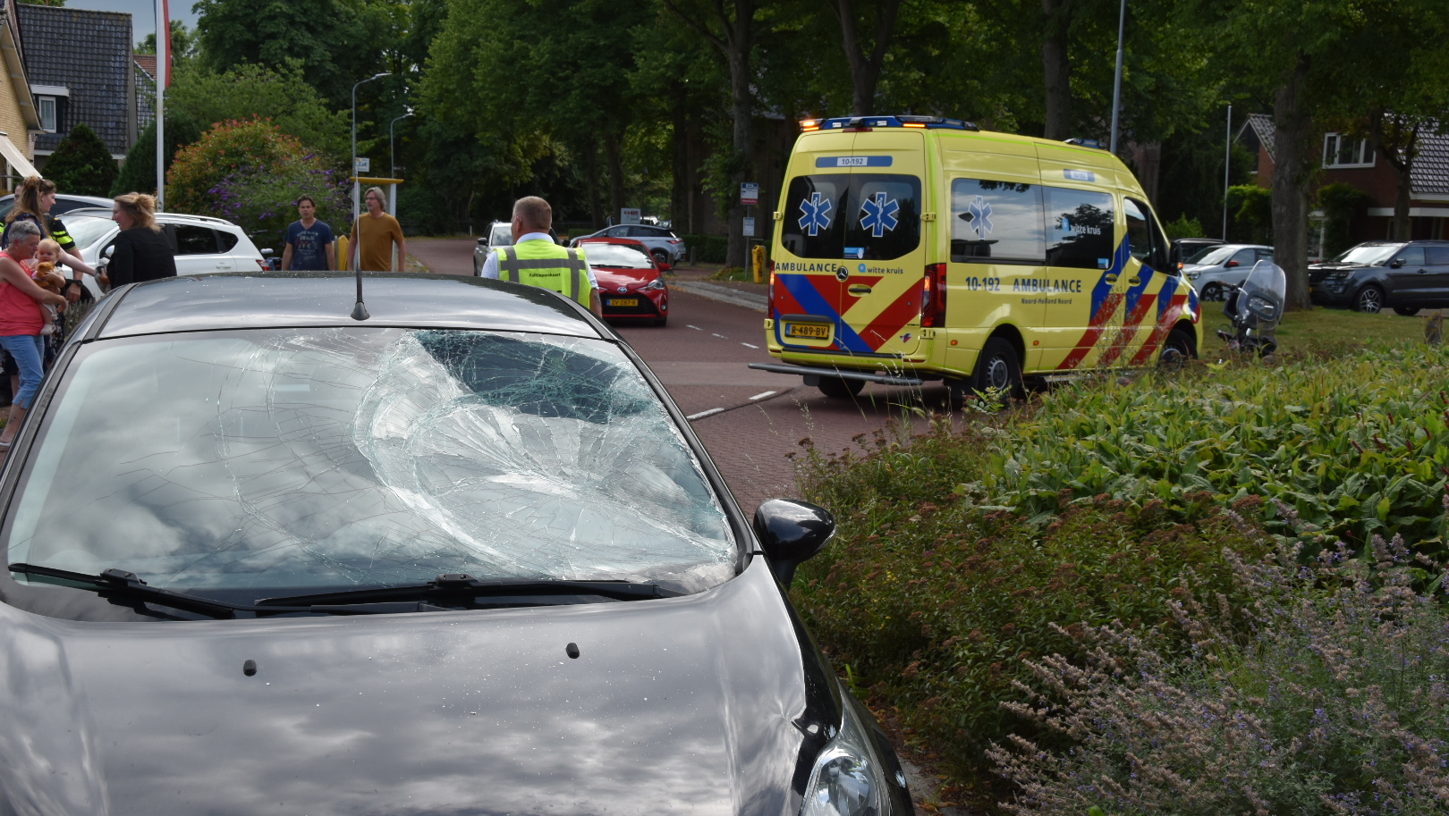 Auto met gebarsten voorruit op straat, met een ambulance op de achtergrond en mensen die zich verzamelen op de stoep.