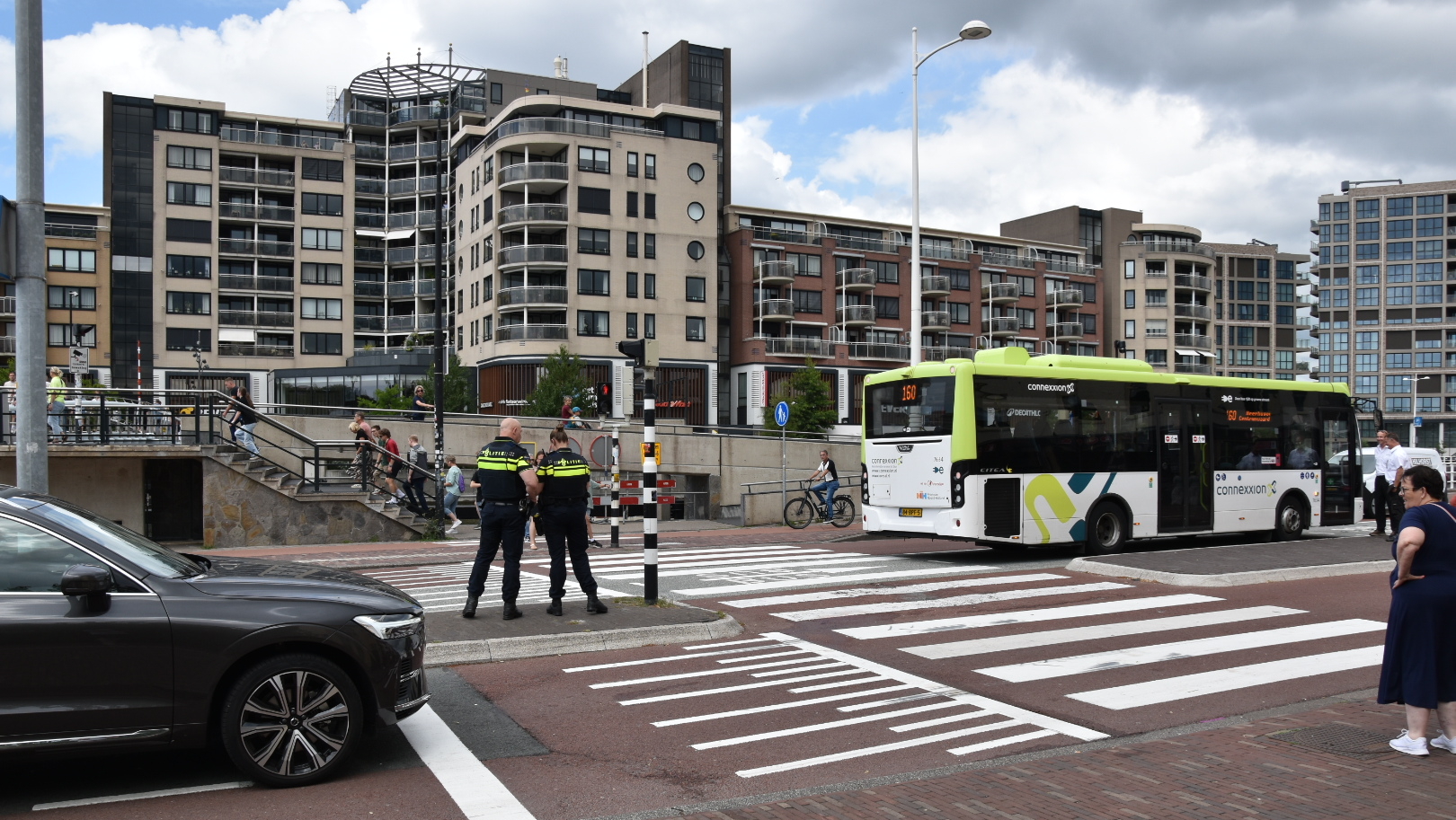 Twee politieagenten staan bij een zebrapad, terwijl een bus en auto's passeren, met een appartementencomplex op de achtergrond.