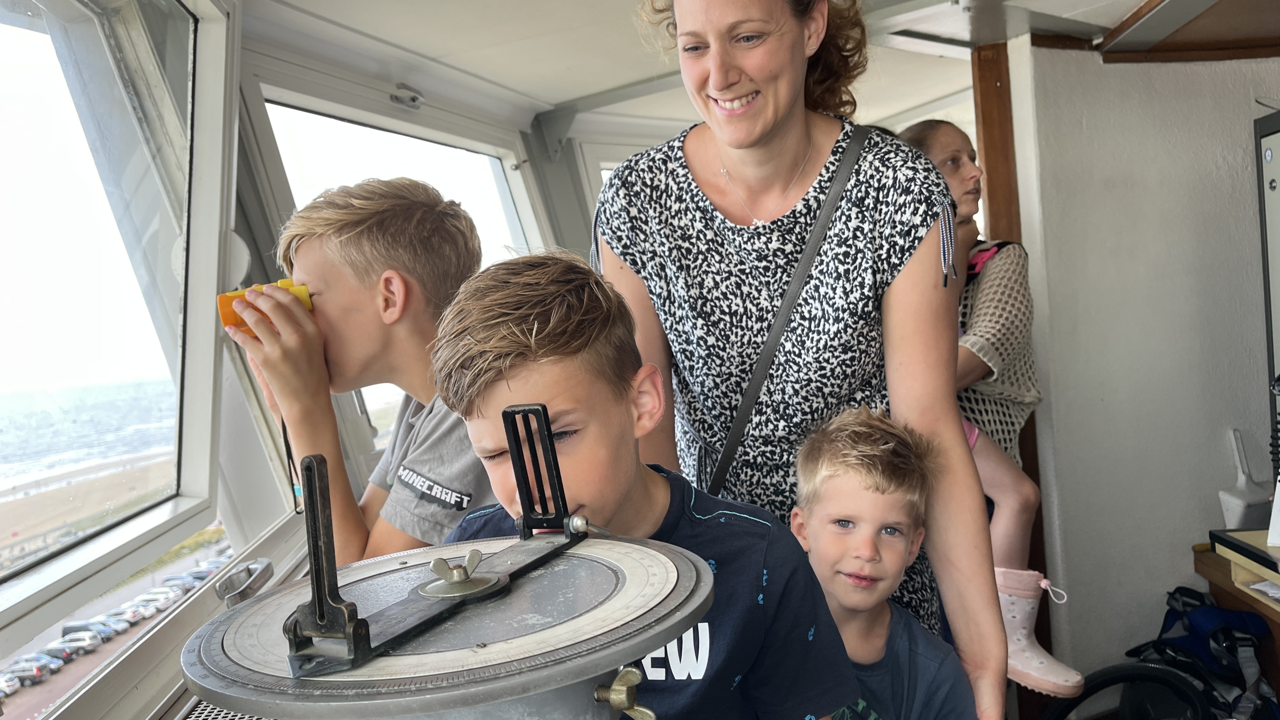 Een glimlachende vrouw staat met drie jonge jongens in een toren, waarvan één jongen door een nautisch instrument kijkt terwijl een andere door een verrekijker kijkt.