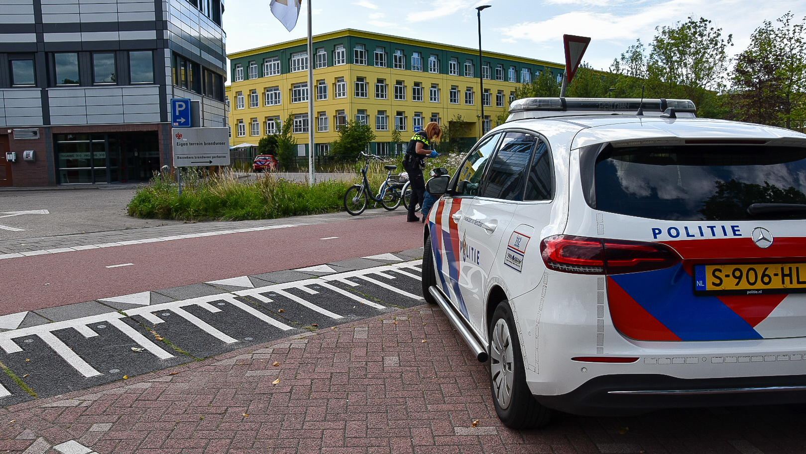 Een Nederlandse politieauto staat geparkeerd op een kruising naast een fietspad. Op de achtergrond is een politieagent te zien die in gesprek is met een fietser. Het gebouw aan de linkerkant heeft een moderne architectuur met metalen panelen, en op de achtergrond staat een geel gebouw met groene details.