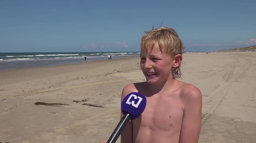 Een jongen met blond haar wordt geïnterviewd op een strand met een blauwe microfoon. Op de achtergrond is de zee zichtbaar onder een heldere blauwe lucht.