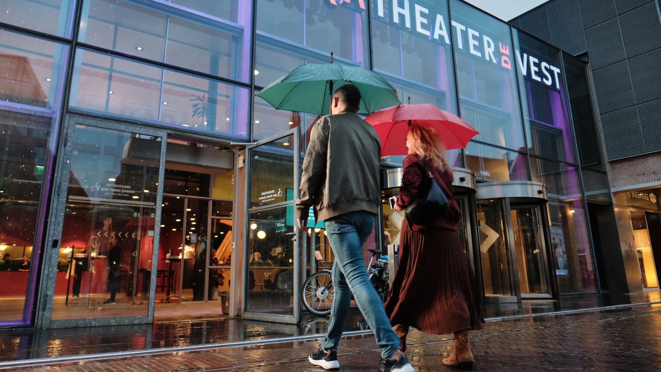 Twee mensen lopen met paraplu's voor de ingang van het TAQA Theater De Vest in de regen.