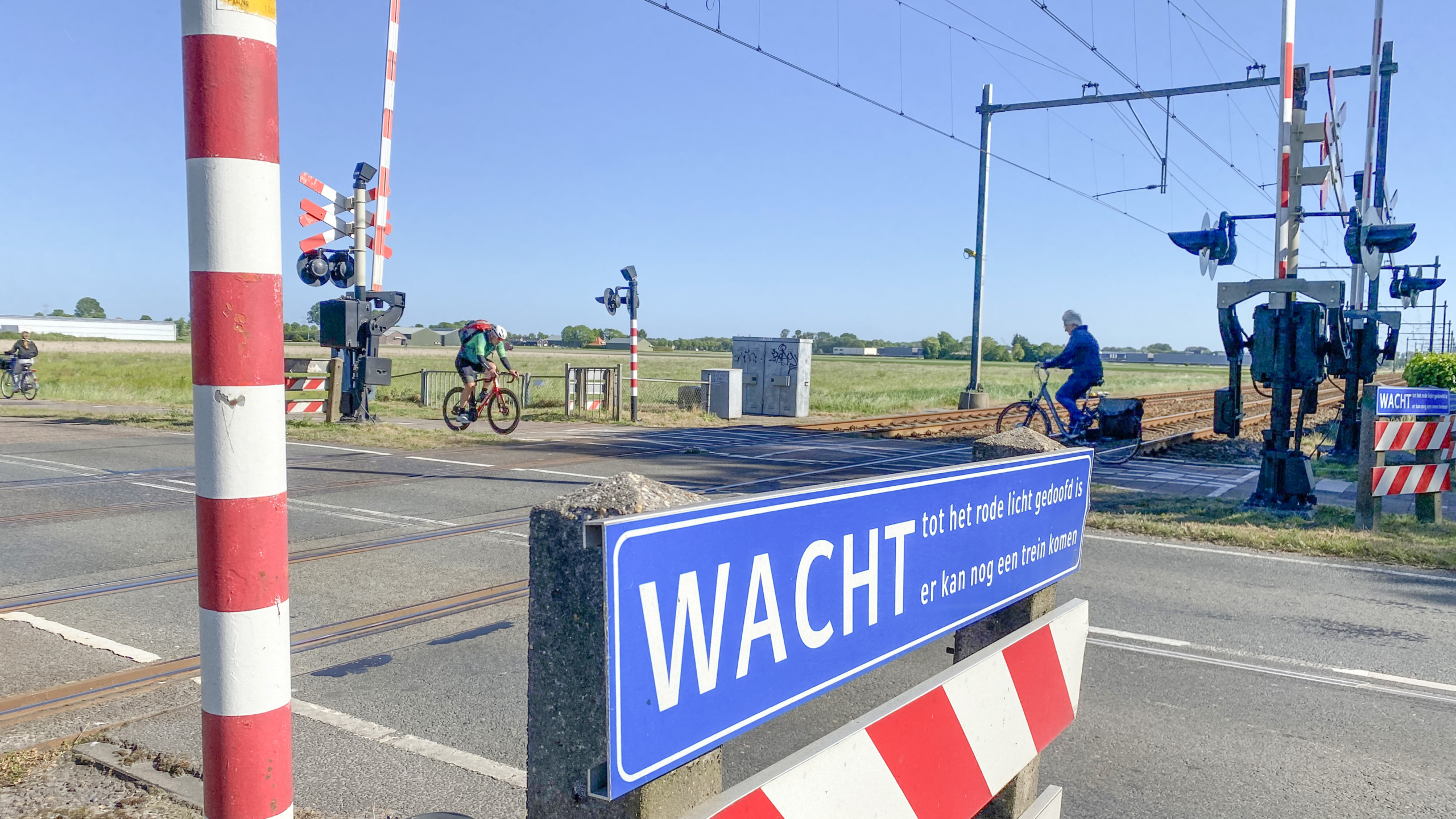 Spoorwegovergang met mensen op fietsen die oversteken, met een bord dat zegt 'WACHT'.