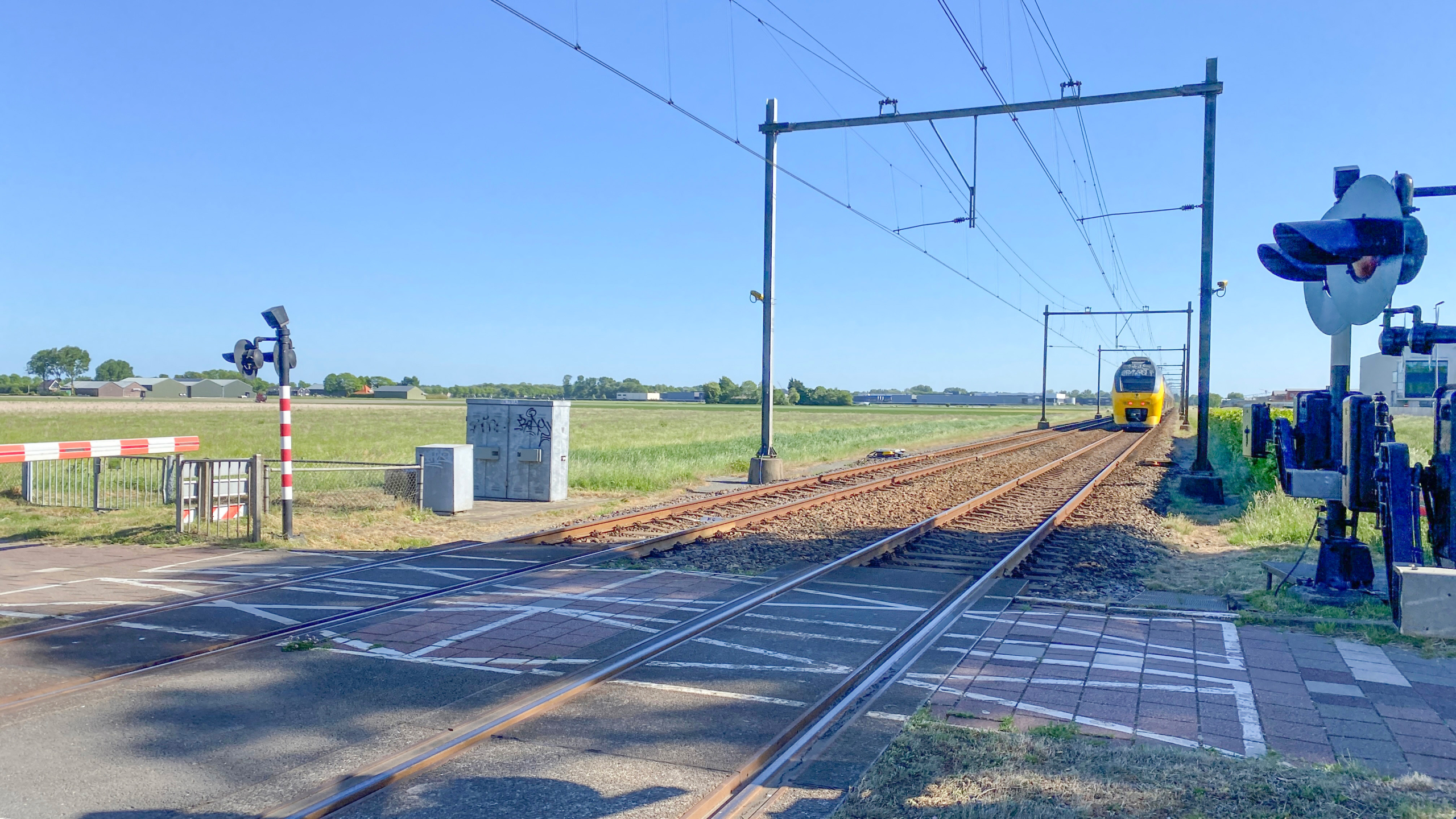 Een geel passagierstrein nadert een spoorwegovergang op een zonnige dag met een open, landelijk landschap op de achtergrond.