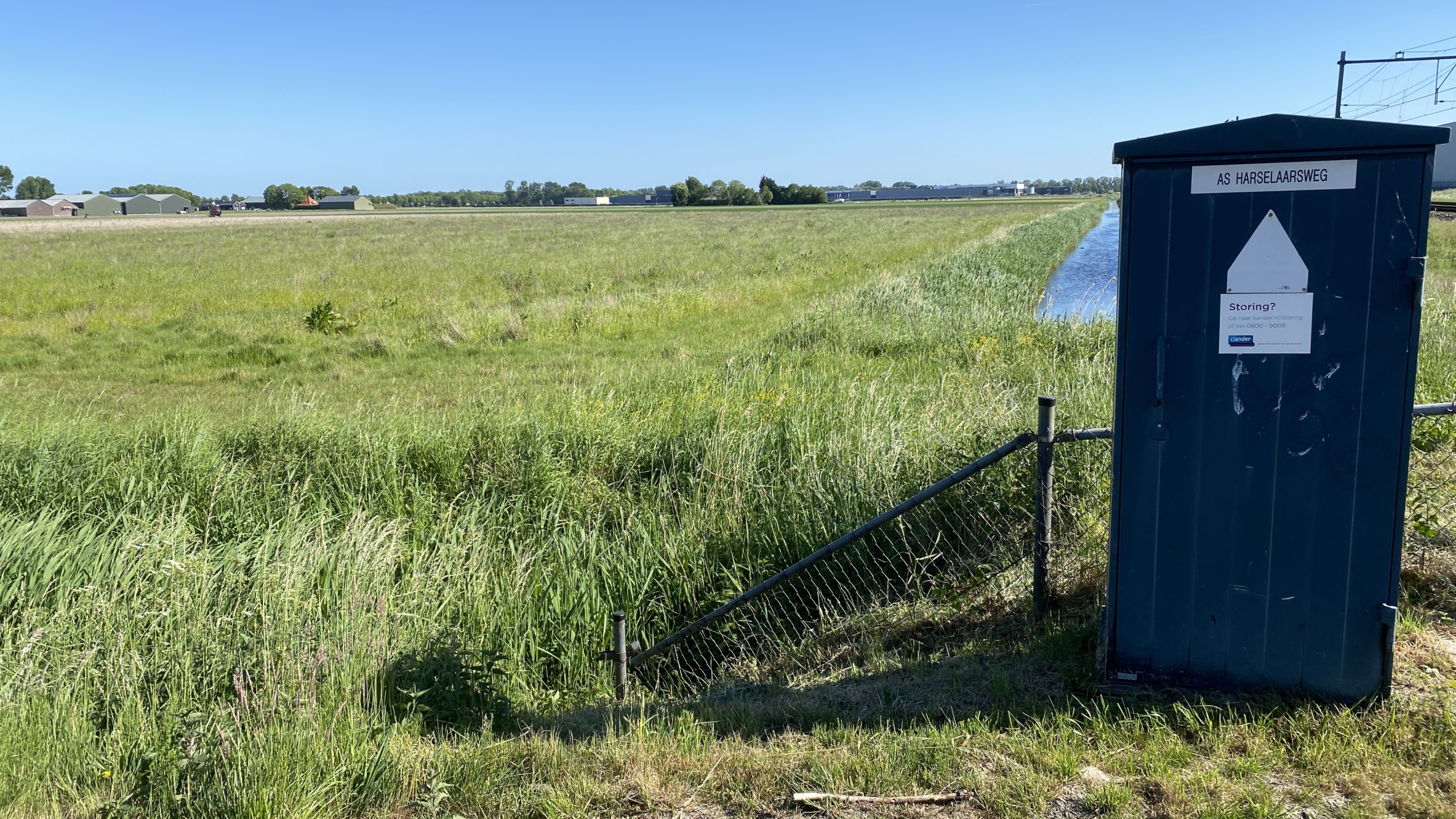 Landschap met een groen grasveld en een sloot rechts, met op de voorgrond een blauw technisch kastje met het label 'AS HARSELAARSWEG'.