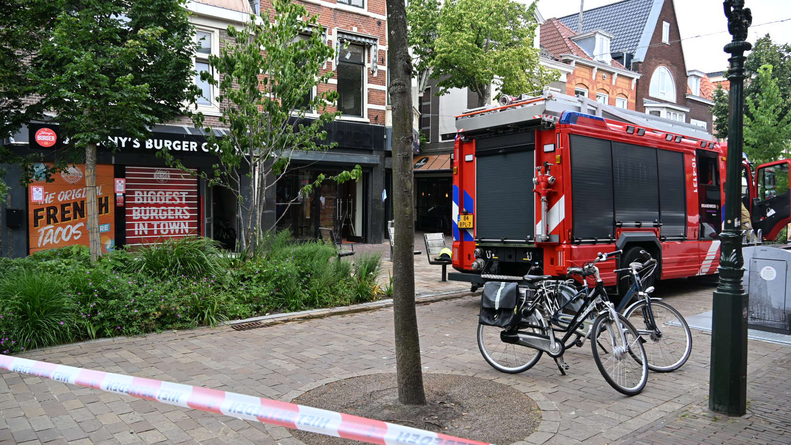 Een rode brandweerwagen staat geparkeerd voor een restaurant genaamd "Marny’s Burger Co.". Het gebied is afgezet met waarschuwingslint, en er staan fietsen geparkeerd naast de wagen. Er is groen gras en bomen in de omgeving.