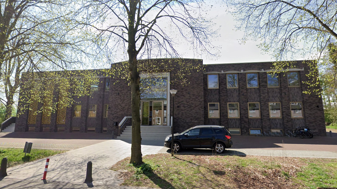 Een donkerbruin, modern gebouw met meerdere ramen en een centrale ingang, geflankeerd door bomen en een geparkeerde zwarte auto op de voorgrond.