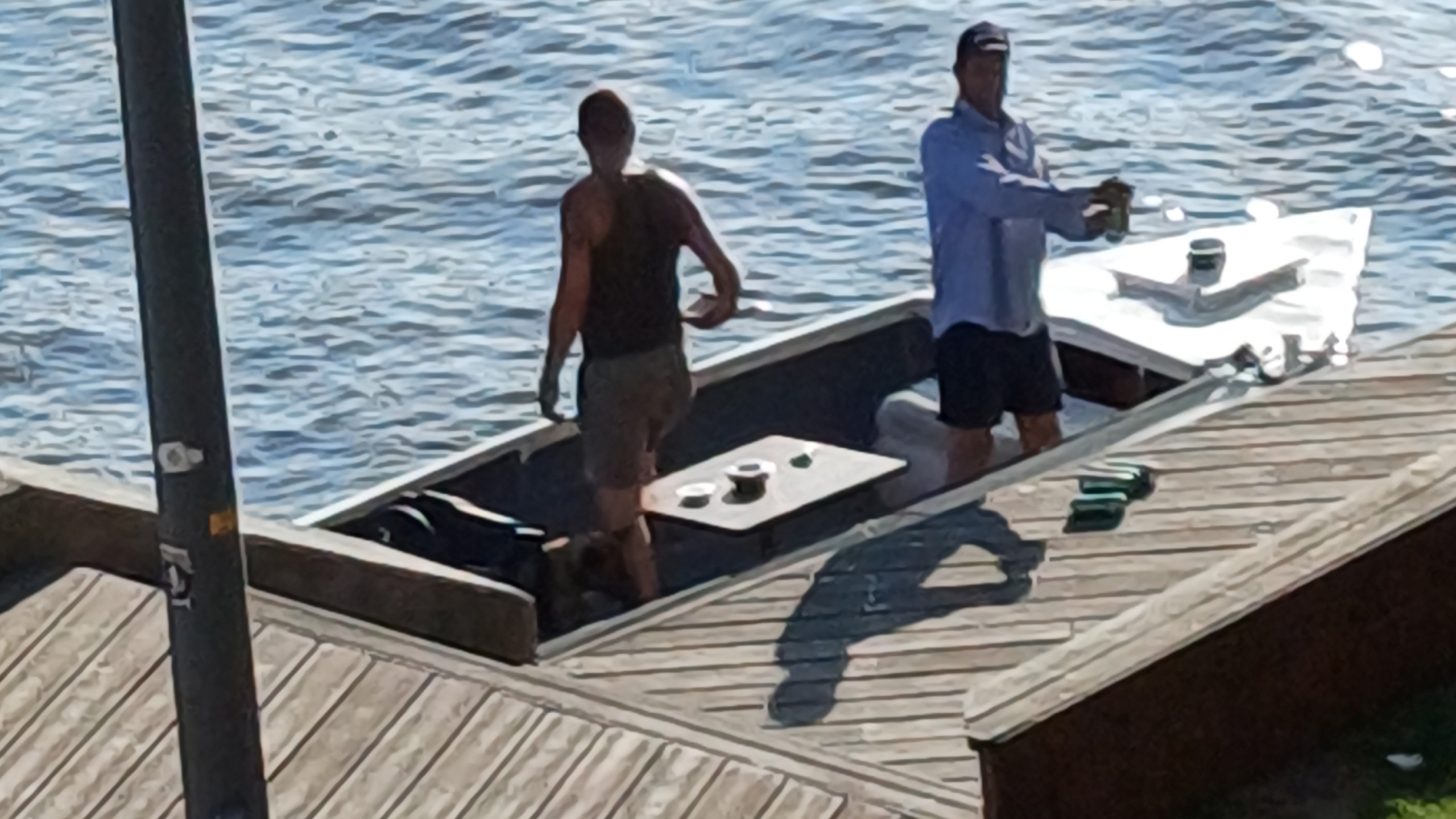 Twee mannen staan op een aanlegsteiger naast een boot op een meer, omringd door water. Een man draagt een tanktop en korte broek, de andere draagt een shirt met lange mouwen en een pet. Er liggen spullen op de steiger en in de boot.