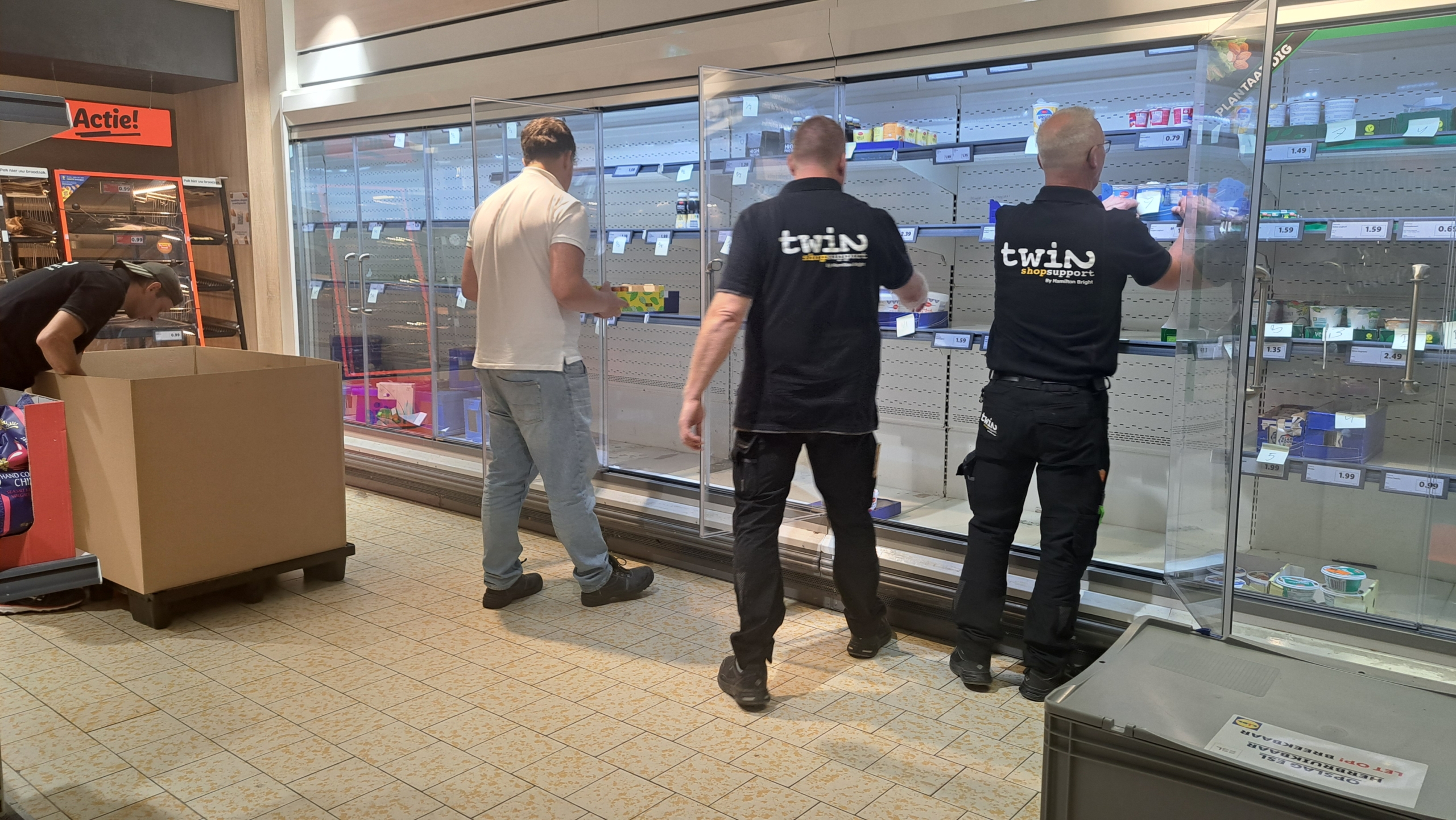 Mensen vullen lege schappen van een supermarktkoeling aan, met een kartonnen doos op de voorgrond en een "Actie!"-teken linksboven.