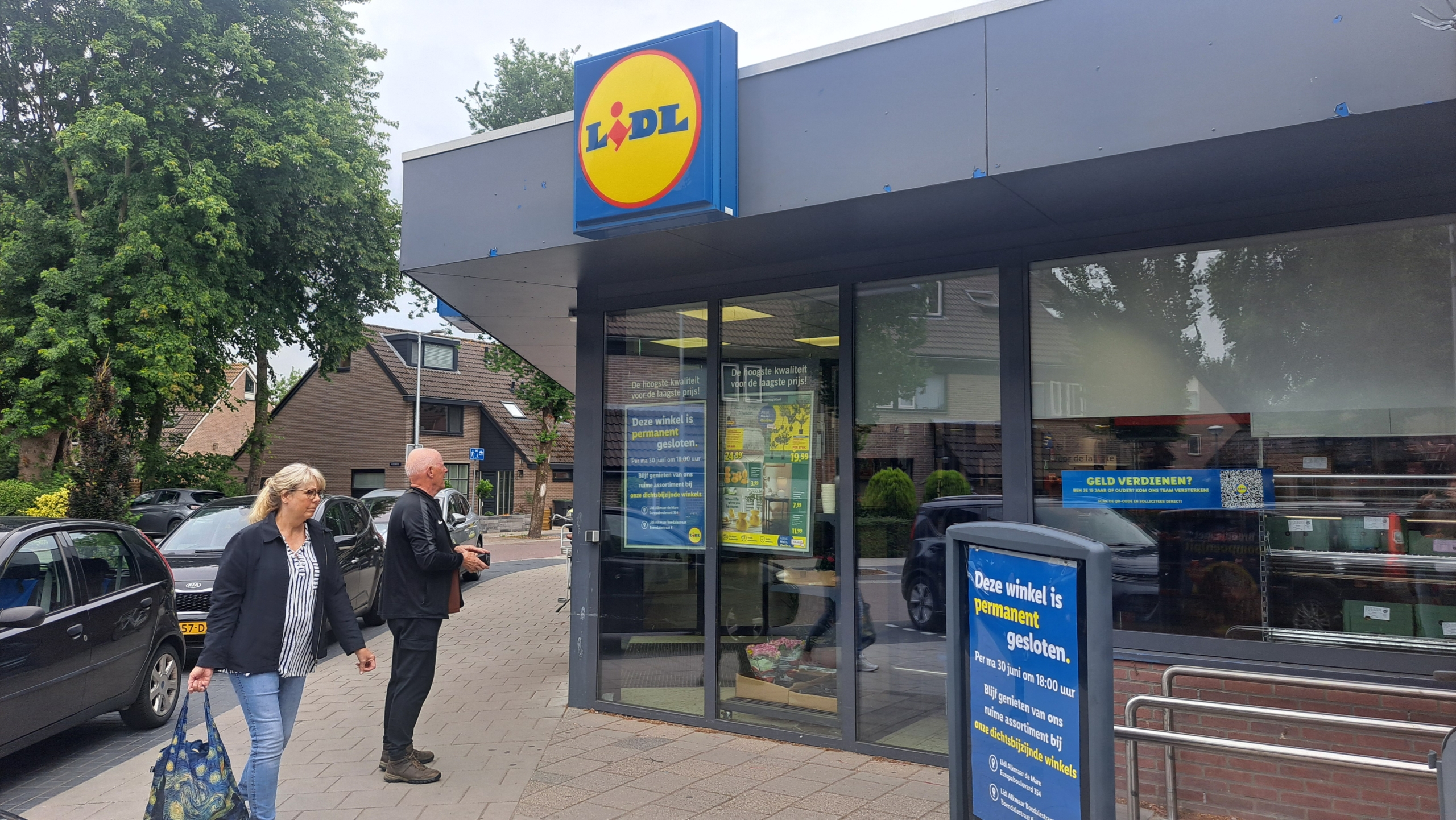 Vooraanzicht van een gesloten Lidl-winkel met borden op de deur die de sluiting aankondigen, naast een voorbijlopende vrouw en een man die richting de winkel kijkt, in een straat met parkeergelegenheid en groene bomen.