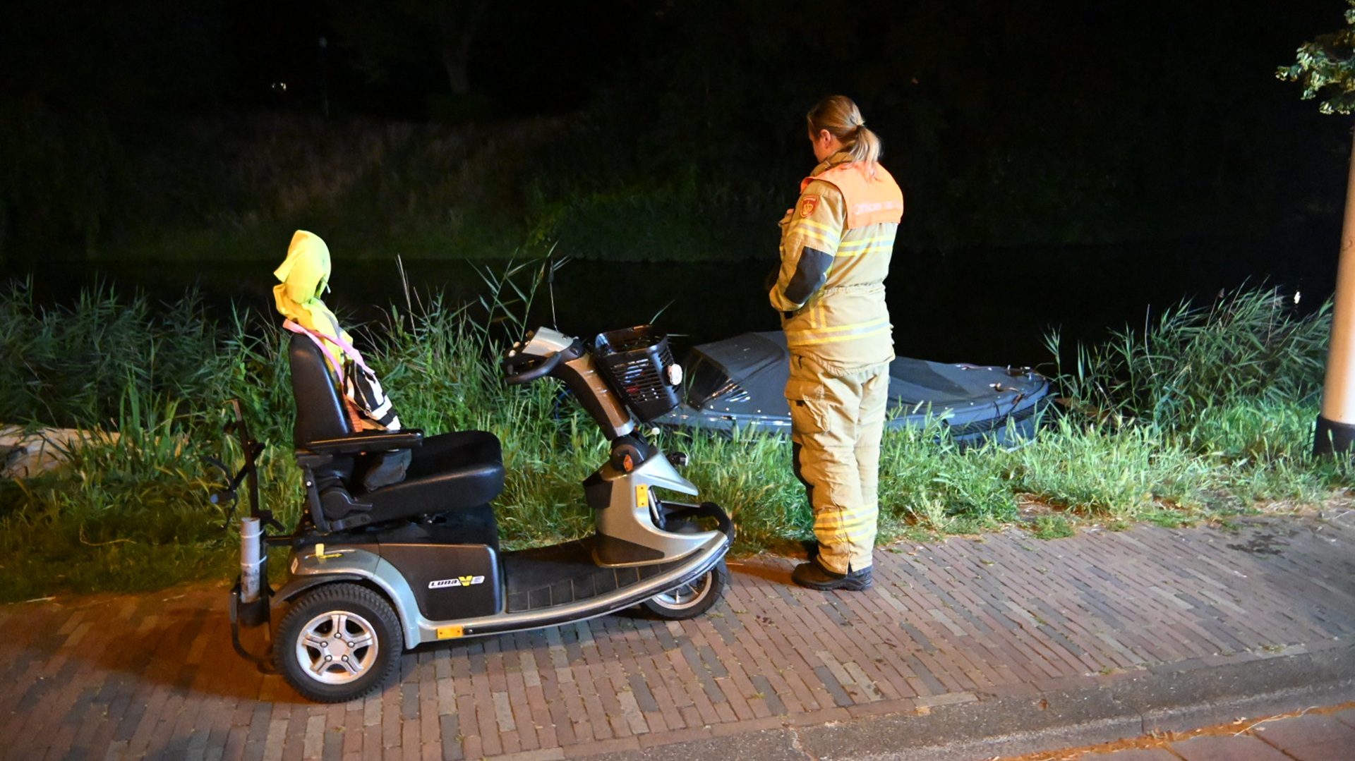 Een brandweerpersoneel staat bij een scootmobiel naast een kanaal in het donker, met een voertuig gedeeltelijk te zien in het water.