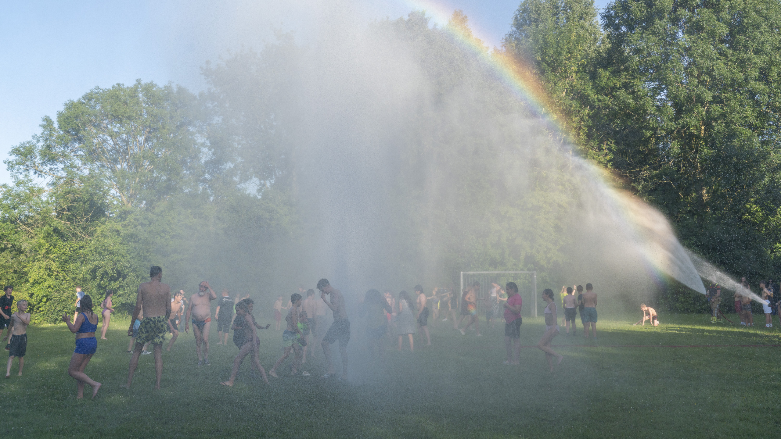 Groep mensen speelt in een park onder een waterstraal, met een regenboog zichtbaar in de mist.