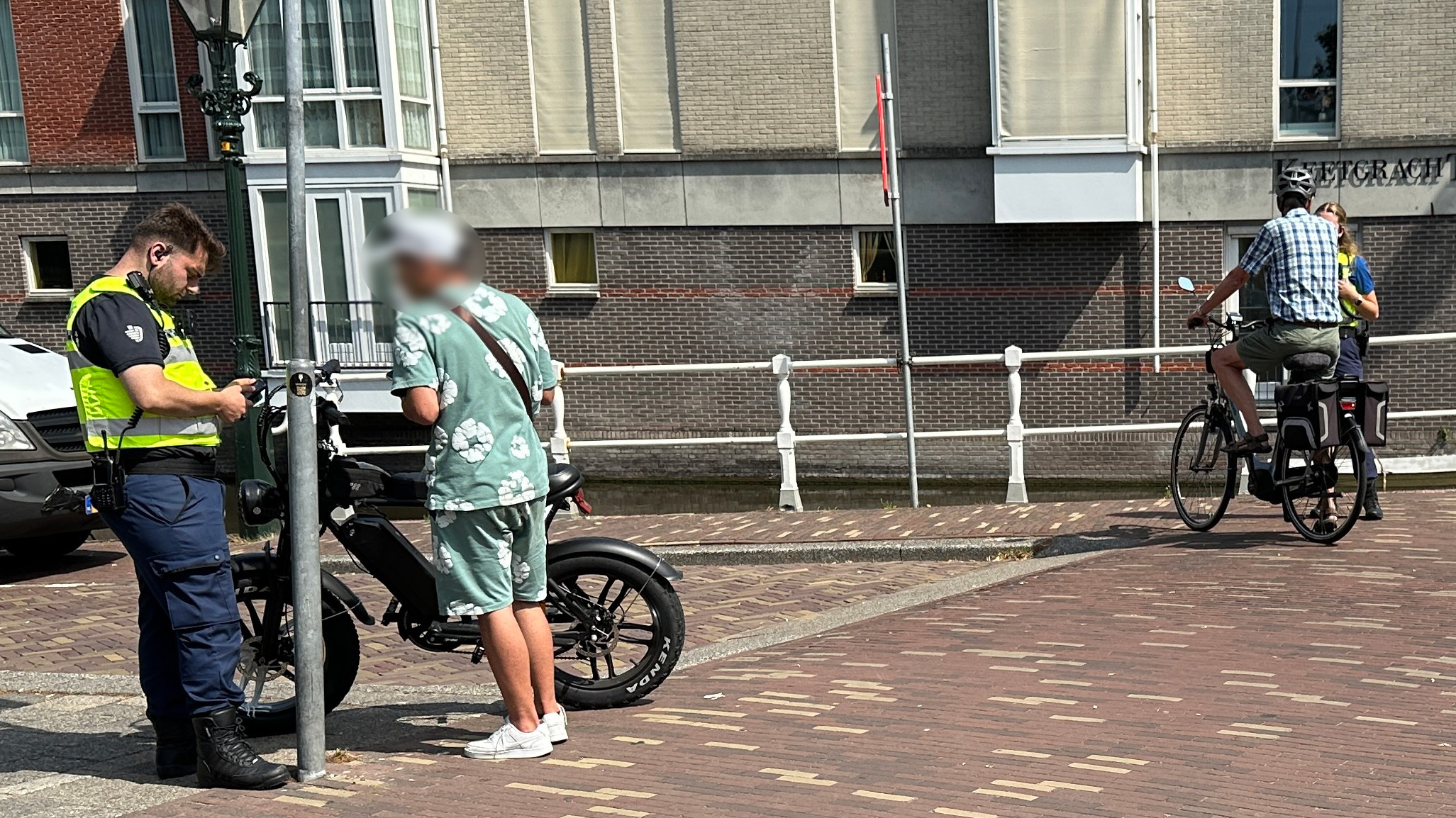 Twee politieagenten in gele veiligheidsvesten zijn in gesprek met mensen op een bakstenen trottoir. Een persoon met een groene outfit staat naast een zwarte elektrische fiets, terwijl een andere persoon op een fiets stilstaat. Op de achtergrond zijn een bakstenen gebouw en een kanaal zichtbaar.