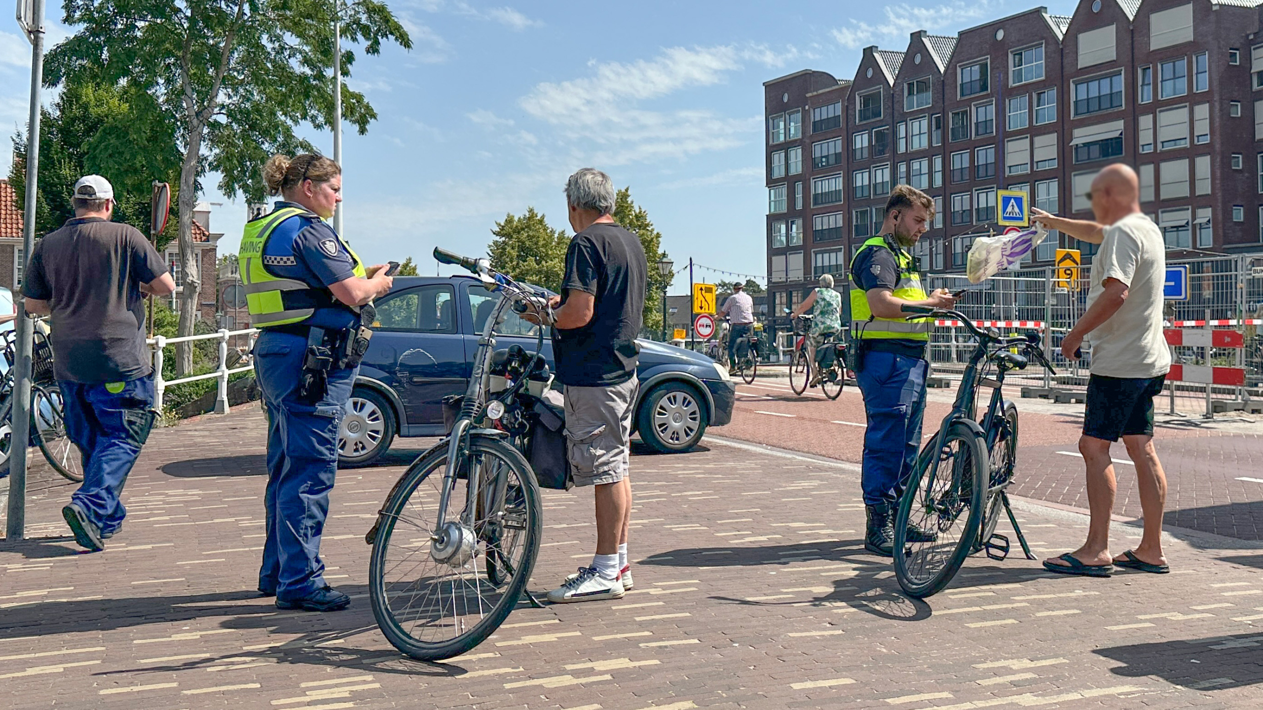Twee handhavers spreken met mannen bij hun fietsen op een zonnige dag; op de achtergrond zijn gebouwen en een paar passerende fietsers te zien.