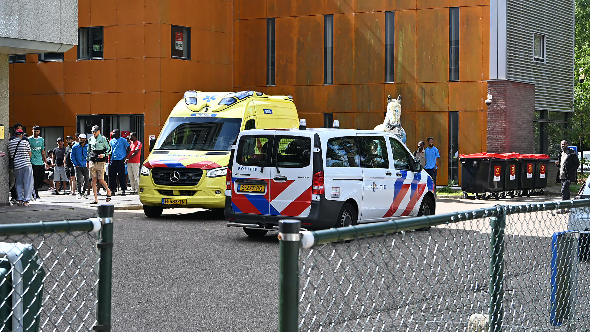 Een politiewagen en een ambulance staan geparkeerd voor een gebouw met een roestbruine gevel, terwijl mensen in de buurt toekijken en langs de zijkant staan.