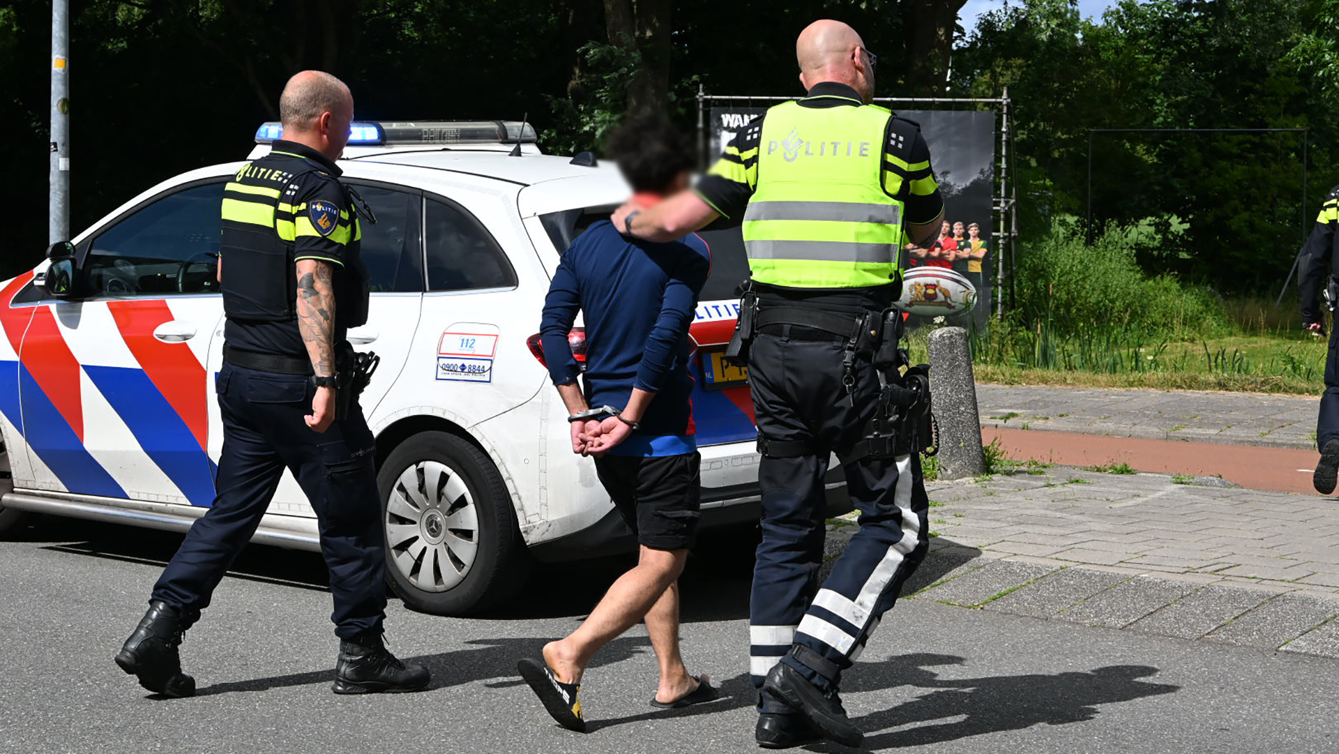 Twee politieagenten escorteren een gearresteerd persoon in handboeien naar een politieauto met blauw en rode strepen.