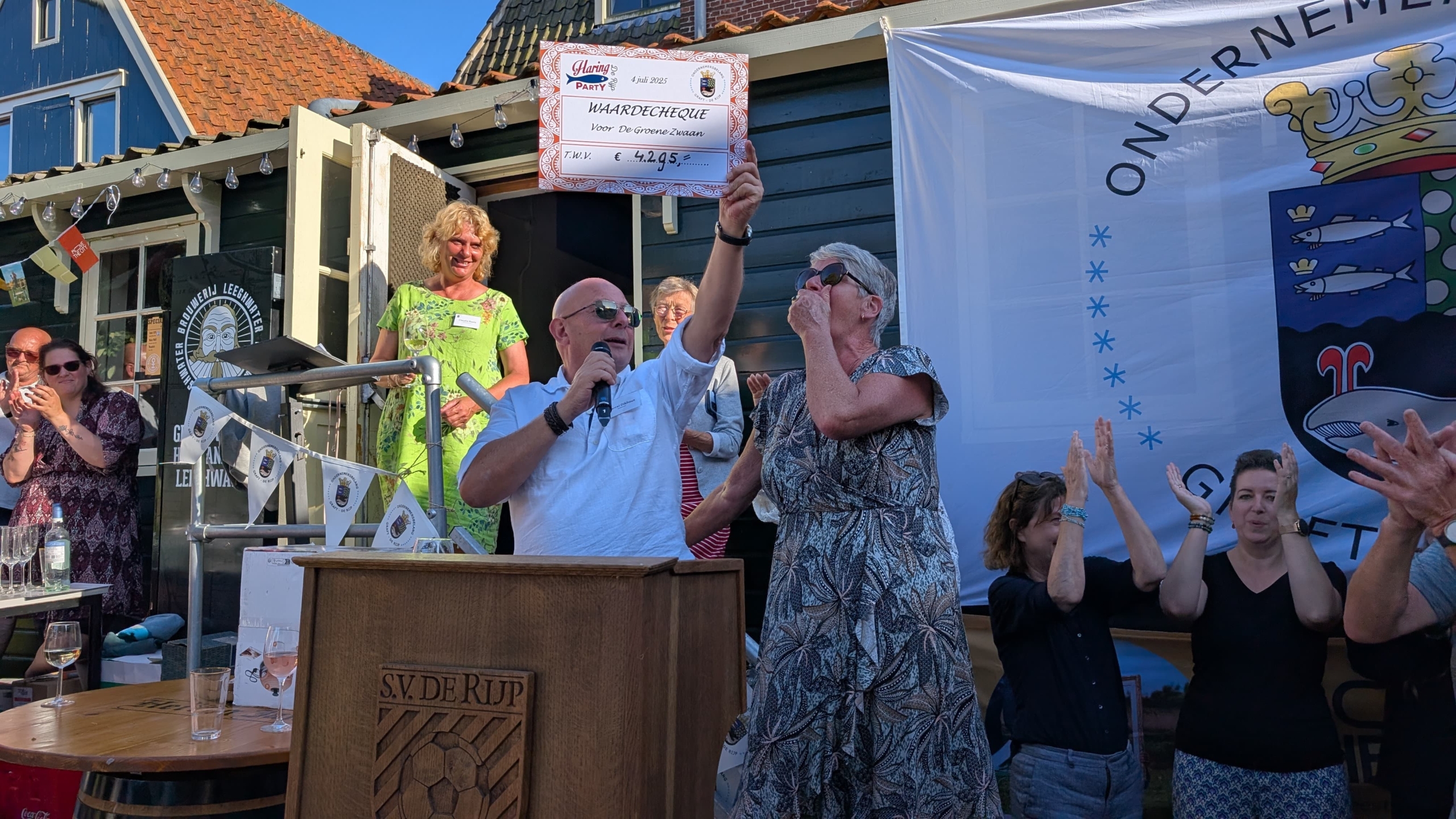 Een vreugdevolle scène van een cheque-overhandiging, waarbij een man met een microfoon en zonnebril een grote cheque omhoog houdt terwijl een vrouw naast hem haar hand voor haar mond houdt van emotie, omringd door klappende mensen in een feestelijke omgeving met een boerderij-achtige achtergrond.