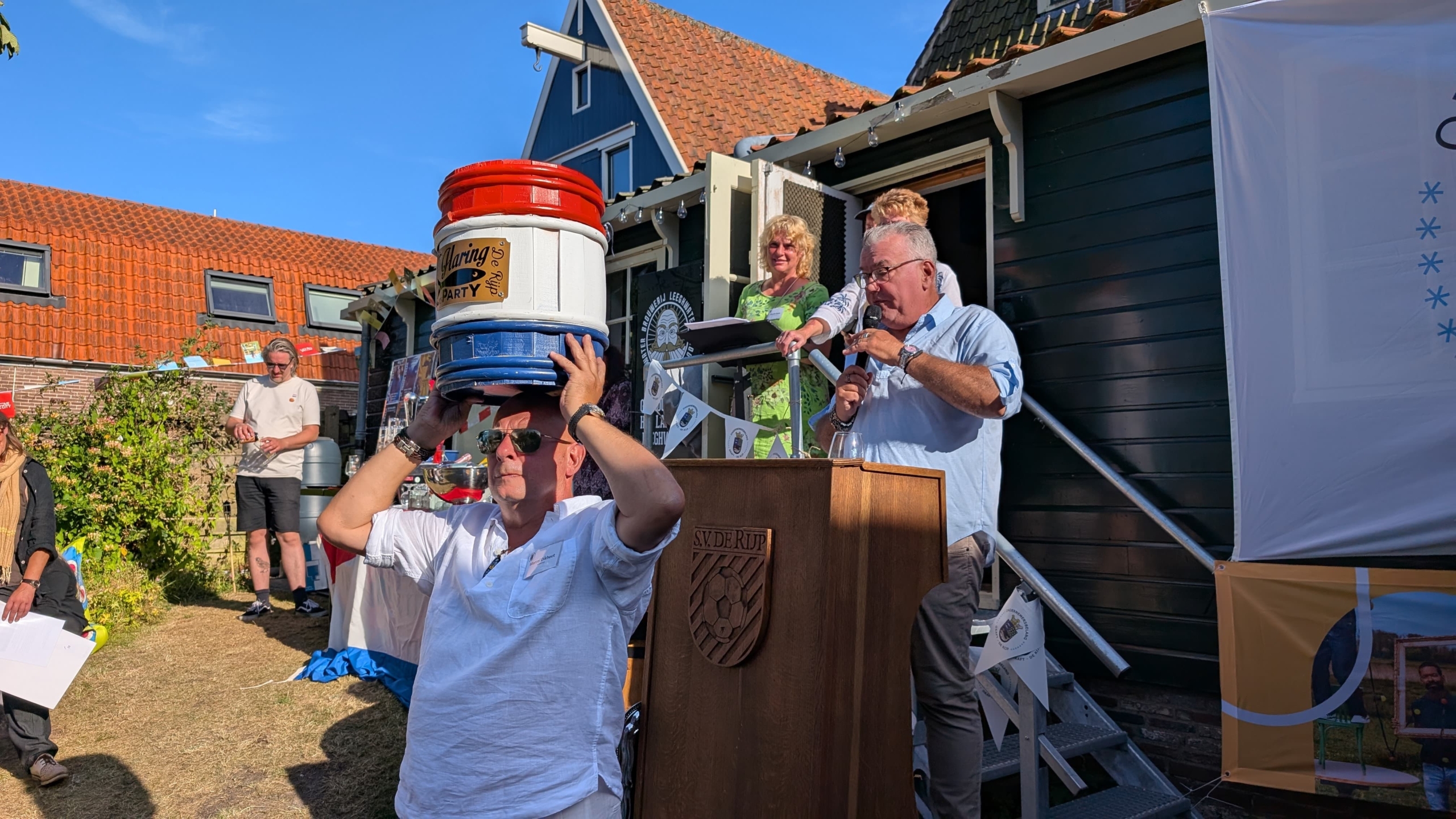 Een man met zonnebril houdt meerdere metalen vaten op zijn hoofd tijdens een buitenbijeenkomst, terwijl een andere man bij een spreekgestoelte in een microfoon spreekt. Op de achtergrond staan mensen en een huis met rood dak onder een blauwe lucht.