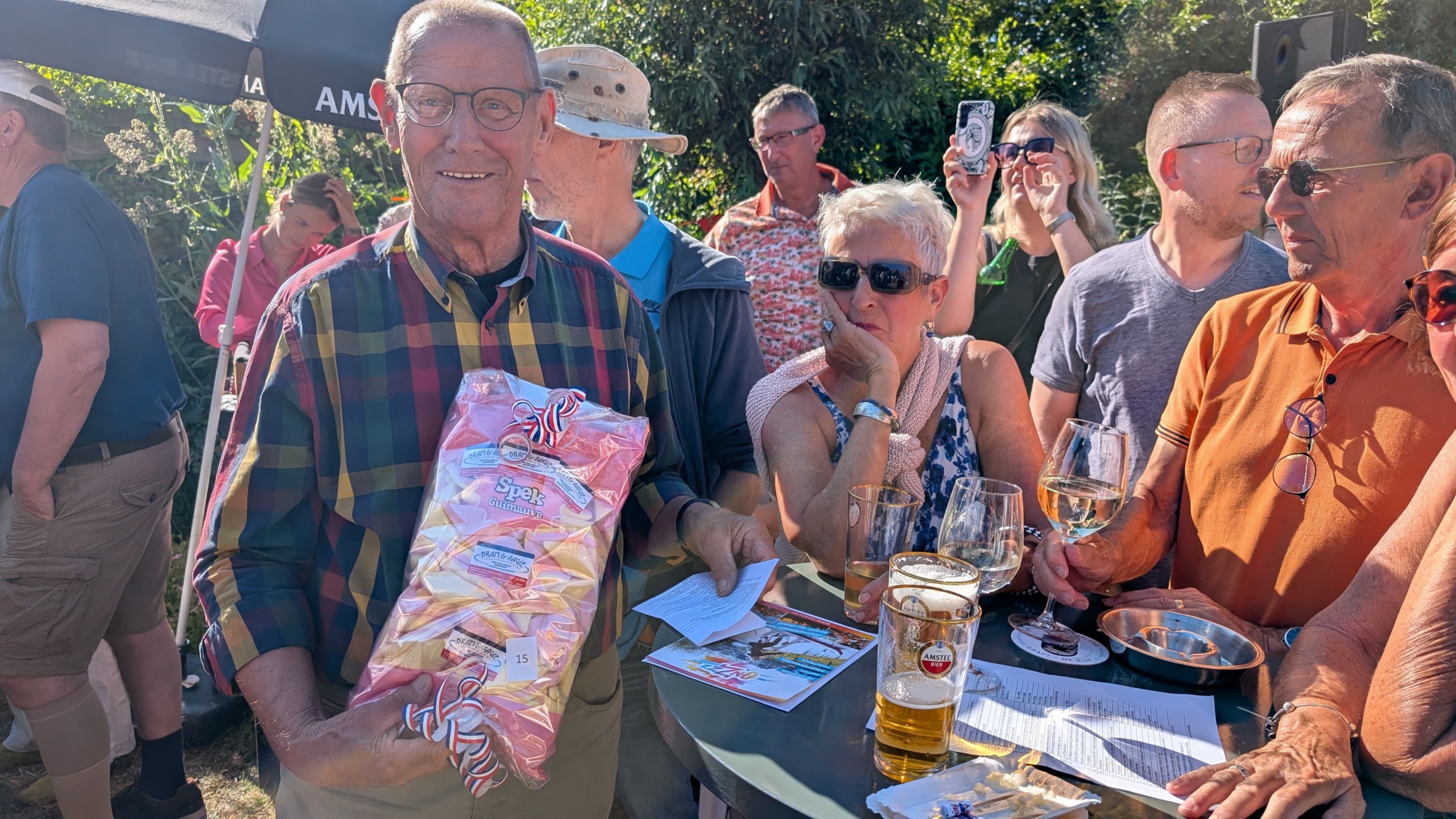 Een oudere man in een geruit overhemd houdt een zak spek snoep vast, omringd door een groep mensen die buiten staan en drinken tijdens een zonnige dag.