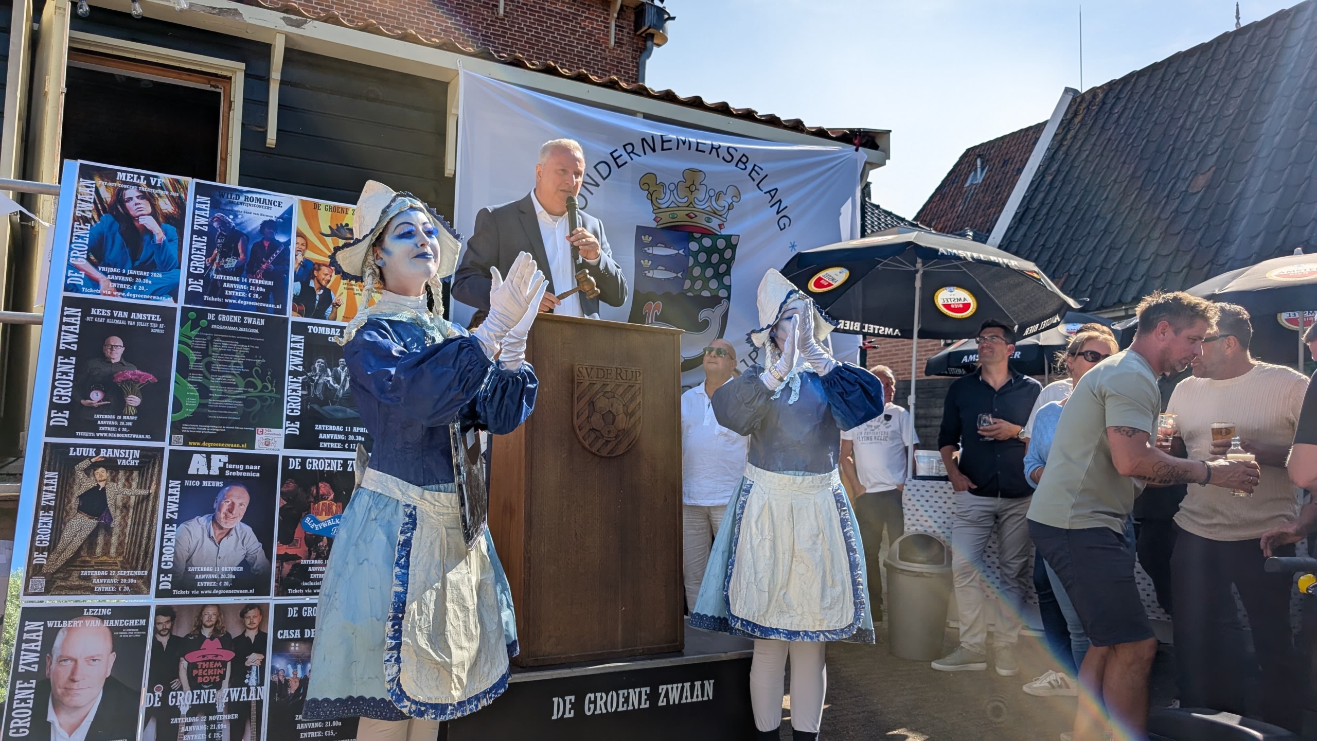 Een man spreekt op een podium met een microfoon, geflankeerd door twee mensen verkleed als poppen in historische kledij. Achter hen is een wand met posters van evenementen te zien en een groep mensen die toekijken. Een vlag met een wapen en de tekst 'Ondernemersbelang' hangt aan de muur. Er zijn ook paraplu's met het logo van Amstel Bier zichtbaar.