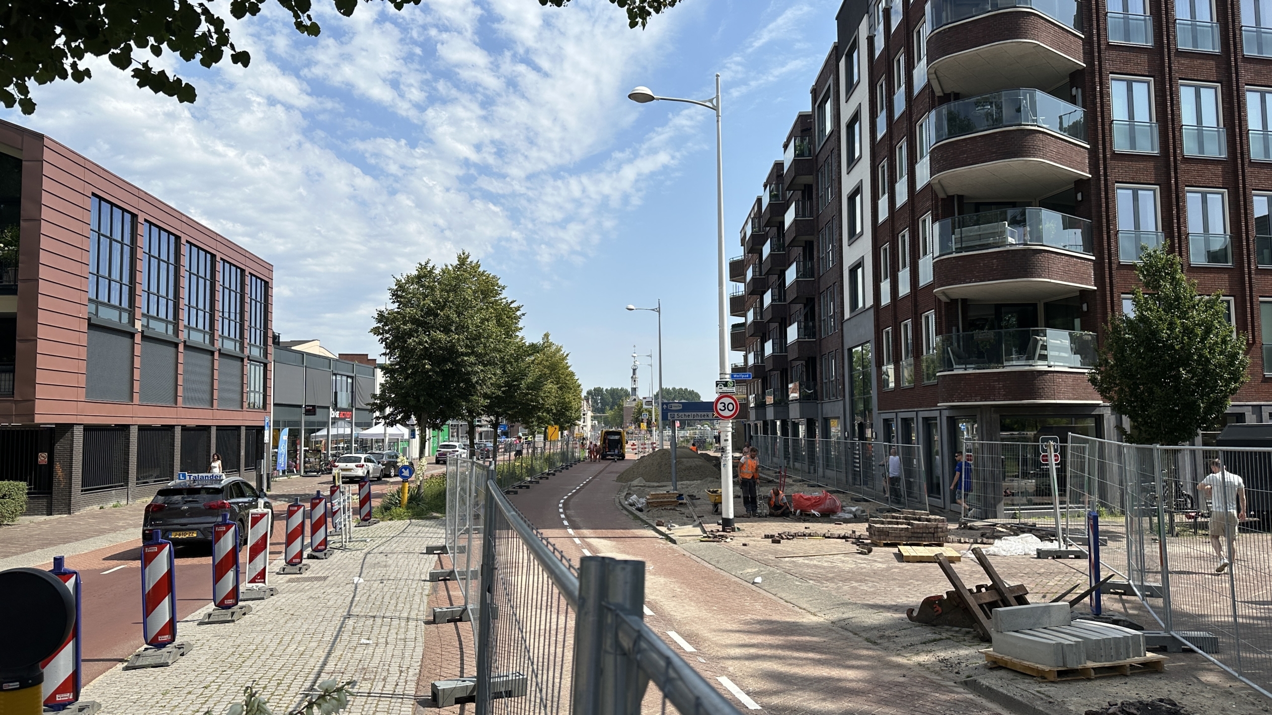 Wegwerkzaamheden aan een straat met moderne gebouwen aan beide zijden, borden voor snelheidsbeperking en diverse bouwwerkzaamheden op de achtergrond, onder een blauwe lucht.