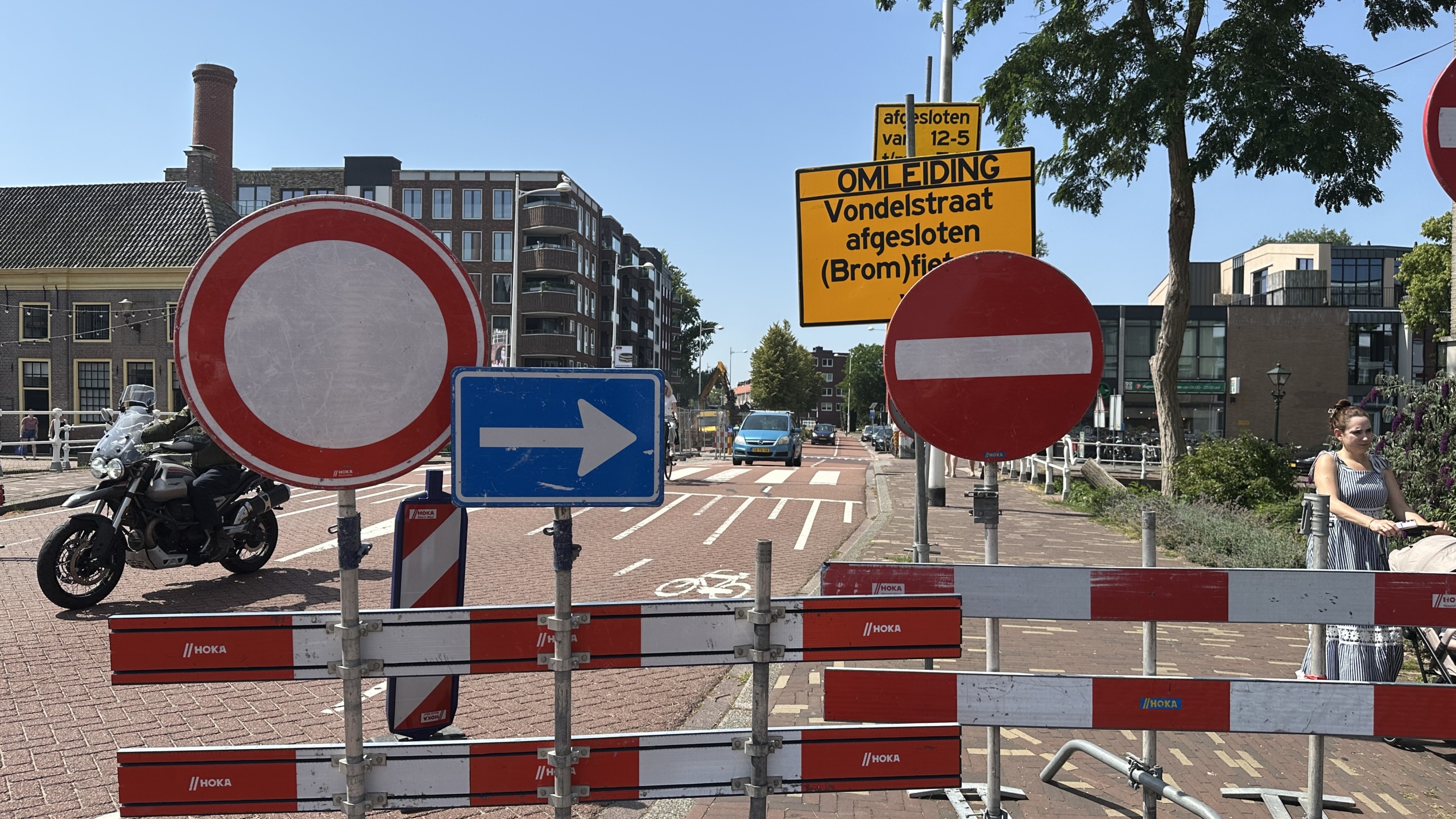 Verkeersborden in een straat met een omleidingsroute aanduiding. Er zijn borden die de weg blokkeren met een blauw bord met een witte pijl naar rechts. Op de achtergrond is een stadsomgeving zichtbaar met een motorfiets en een vrouw die een kinderwagen duwt. Een gele bord geeft aan dat de Vondelstraat afgesloten is voor (brom)fietsers.