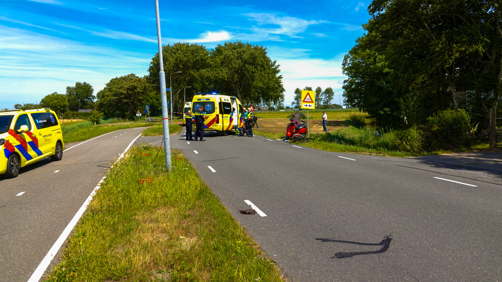 Ambulance en noodvoertuig op een weg bij een verkeersongeluk met een rode scooter aan de kant, omringd door paramedici en een verkeersbord met een waarschuwing voor een kruispunt in de achtergrond.