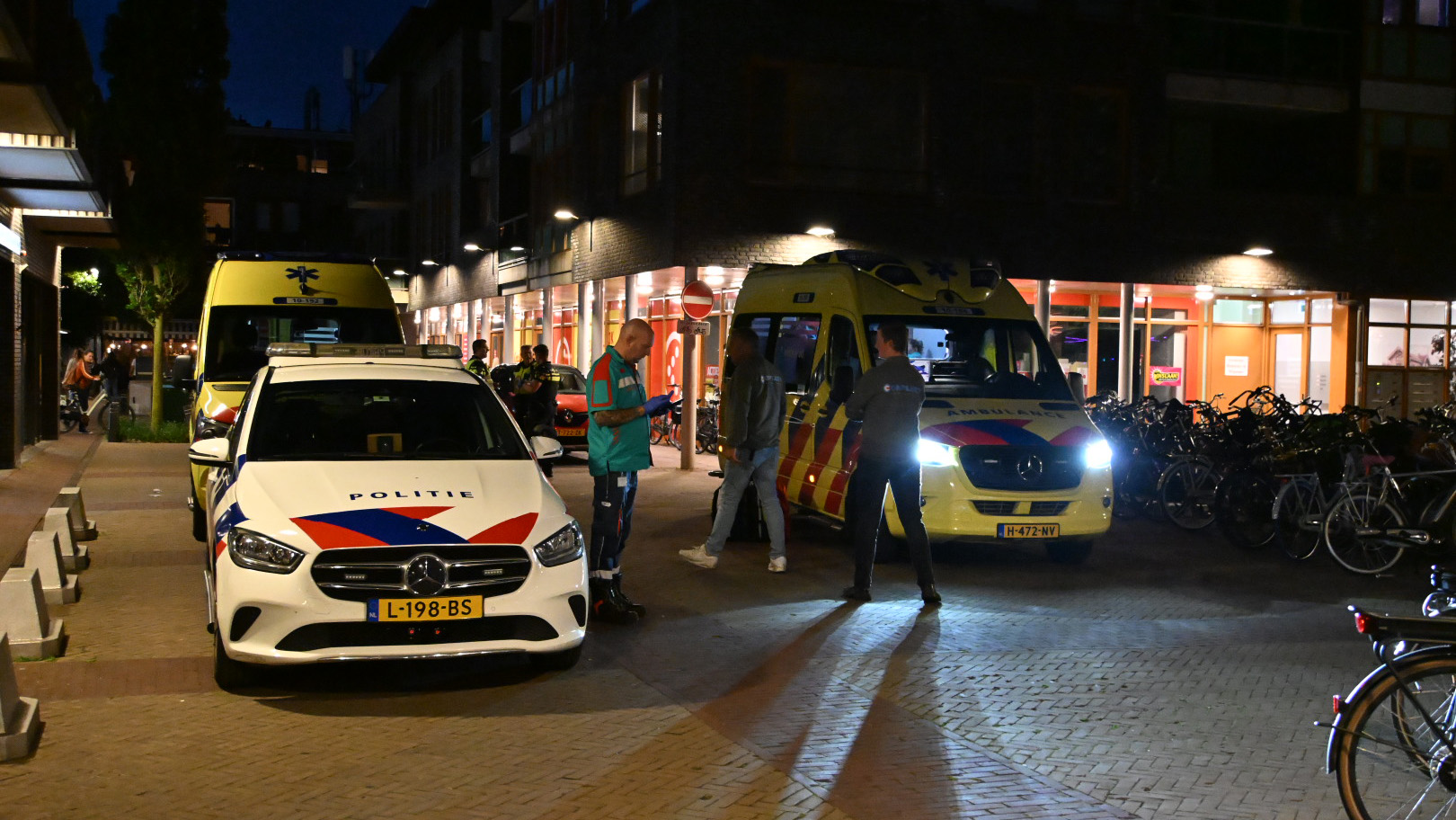 Een politieauto en twee ambulances staan geparkeerd op een straat in de avond, met mensen rondom aanwezigheid, voor een gebouw met fietsenrekken.