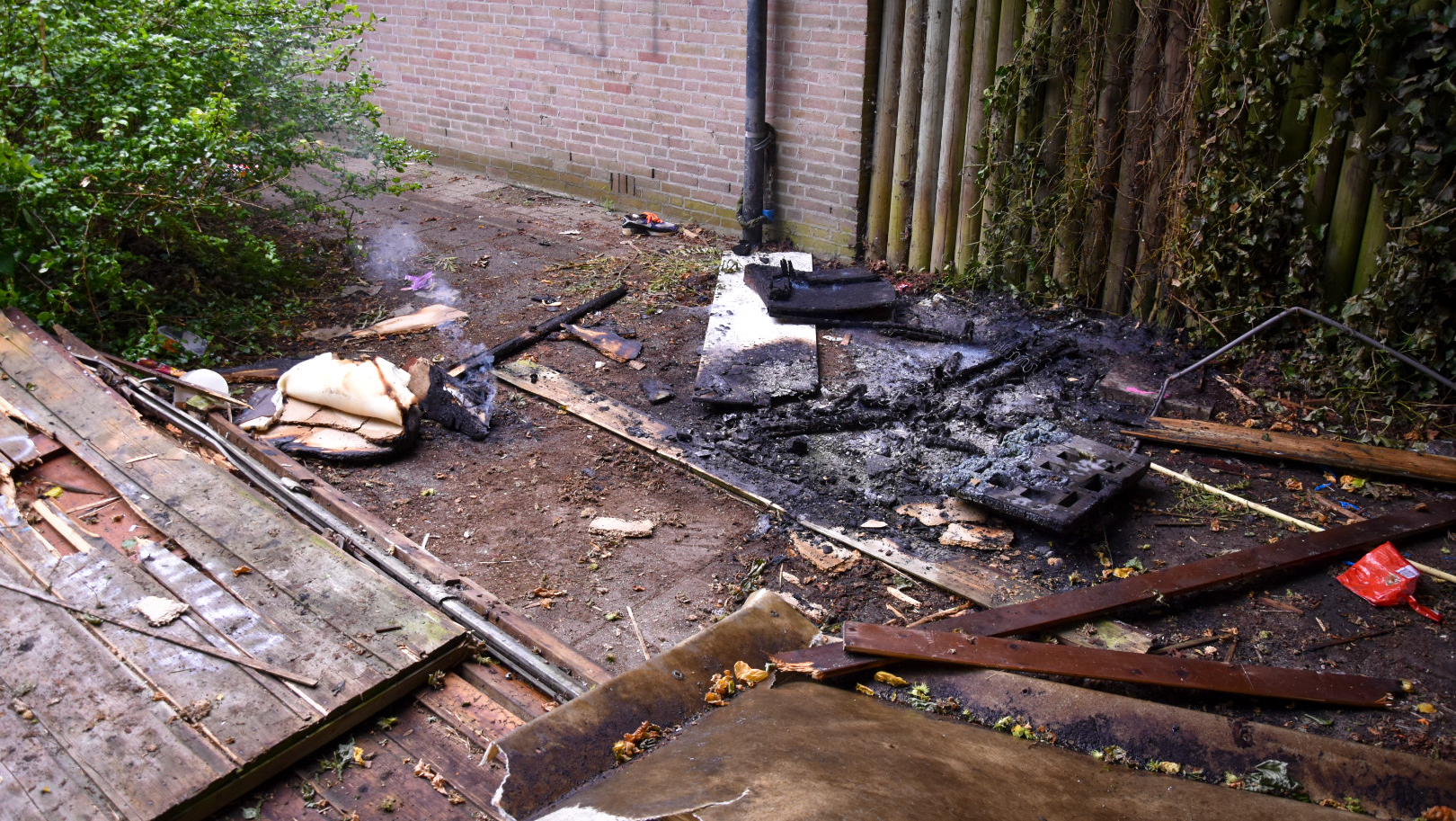 Geblakerde en verkoolde overblijfselen van een brand op de grond naast een stenen muur en een houten omheining, met rook die nog zichtbaar opstijgt, omringd door planten en houtresten.
