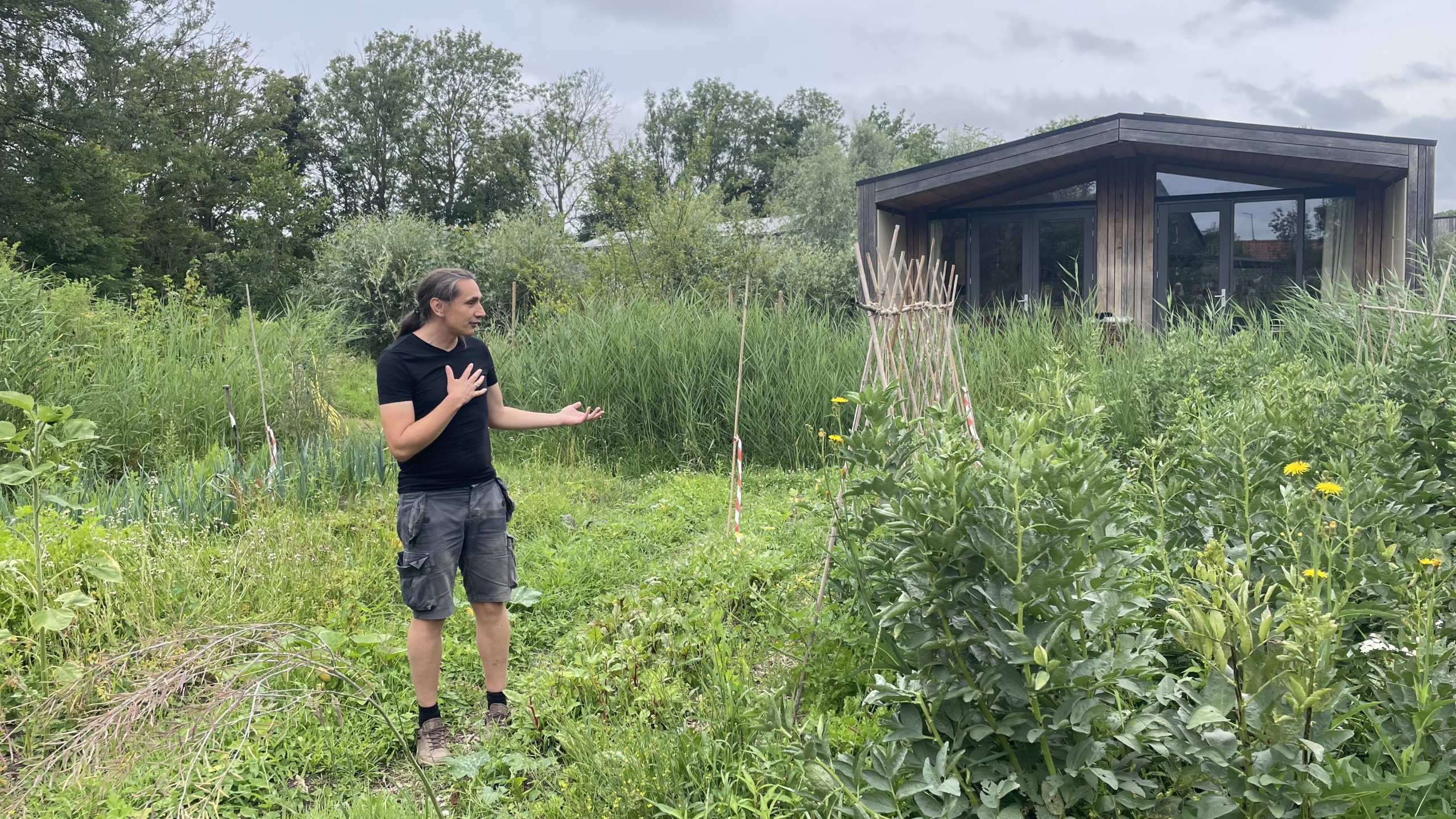 Een persoon in een zwarte outfit staat in een groene tuin met hoge planten en bloemen, naast een houten gebouw.