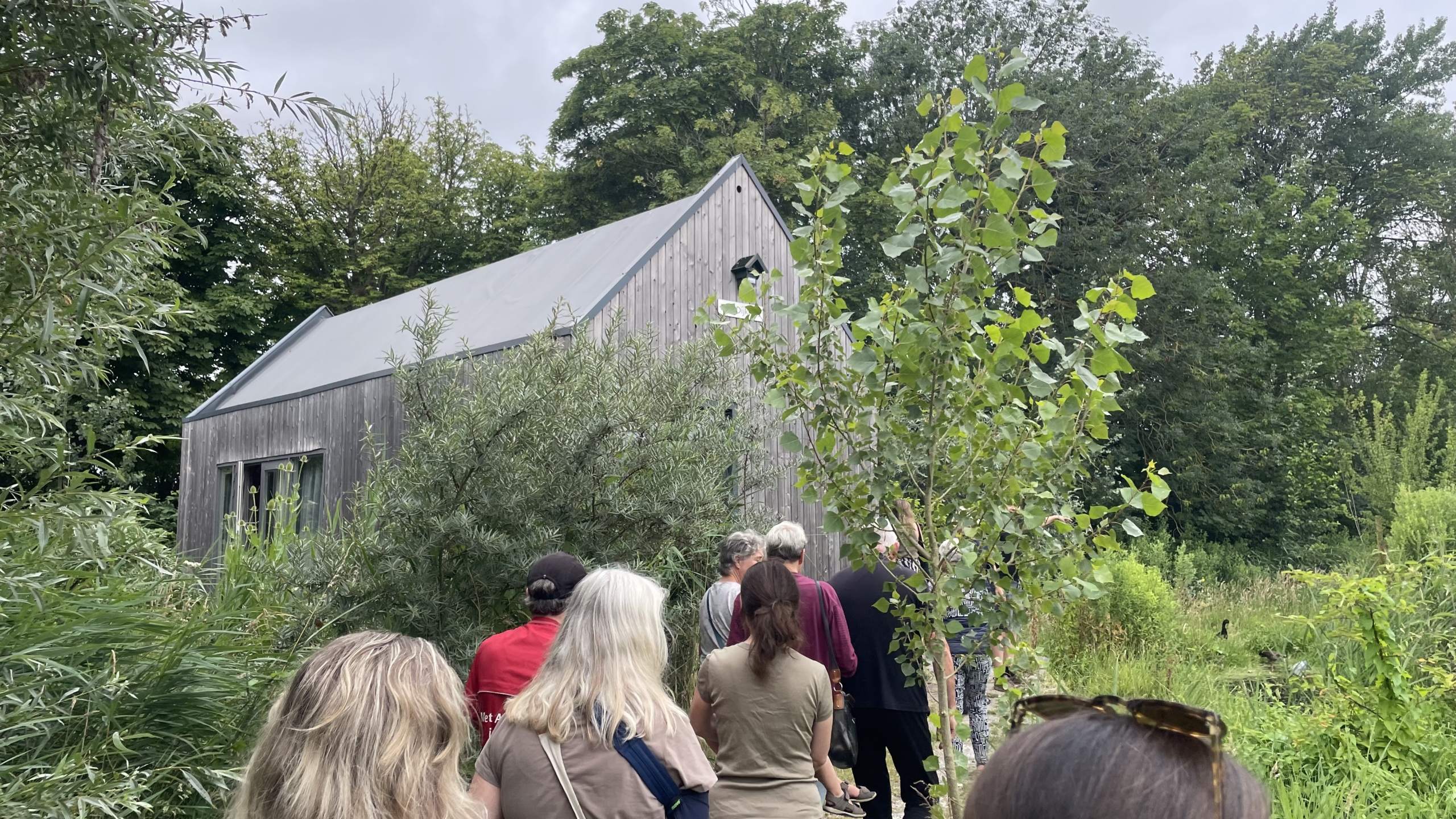 Een groep mensen loopt op een pad door een groene omgeving, naar een houten gebouw dat half verscholen is tussen de bomen en struiken.