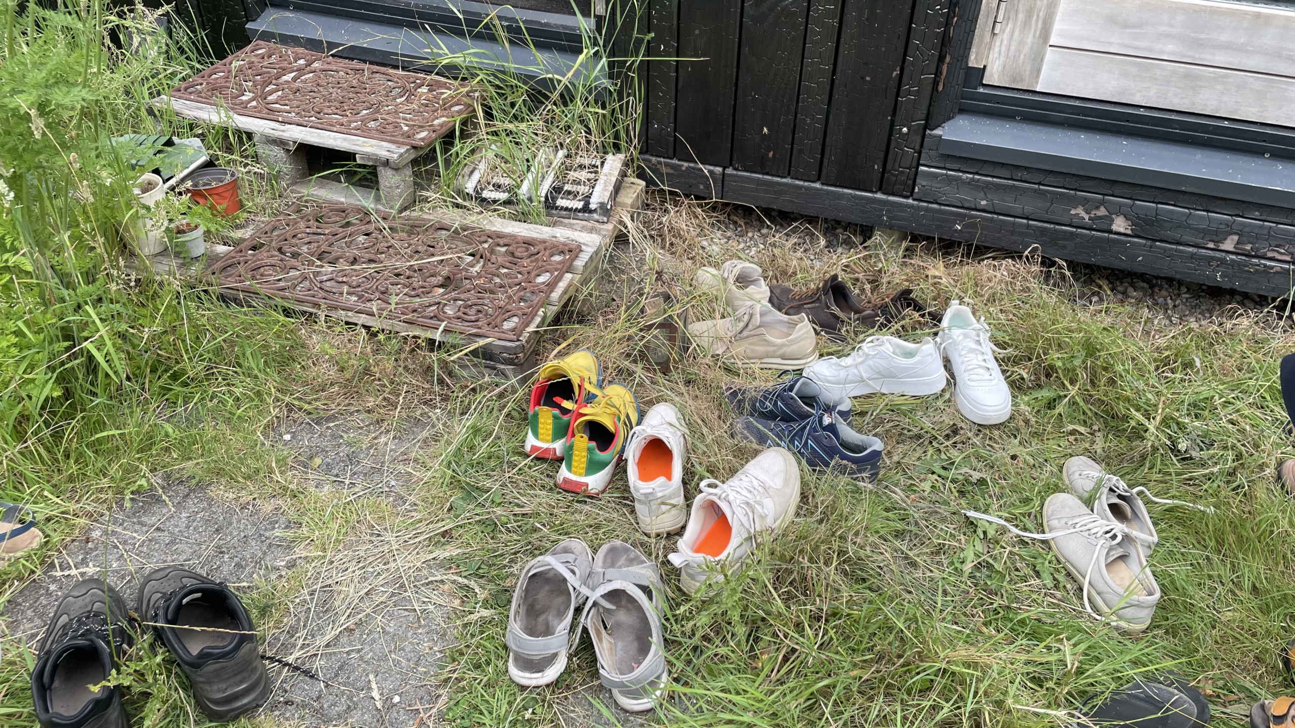 Verschillende paren schoenen liggen verspreid op het gras en een stoepje met decoratieve matten voor een deur, omgeven door wilde planten.