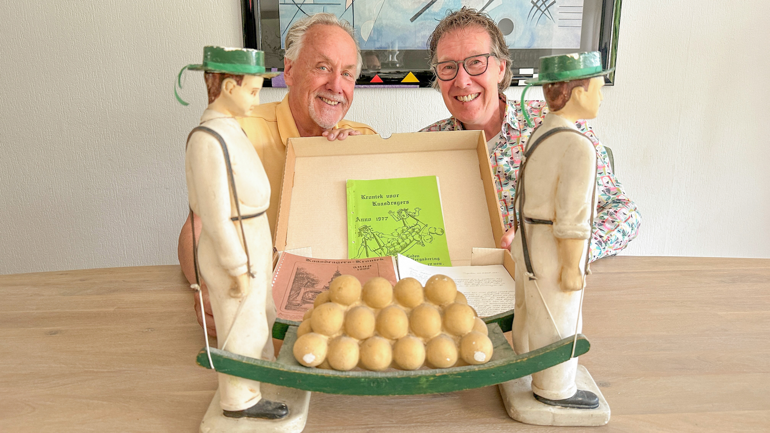 Twee lachende mensen poseren achter een houten beeld van kaasdragers met een doos gevuld met historische kaasdragersboekjes en papieren op de voorgrond.