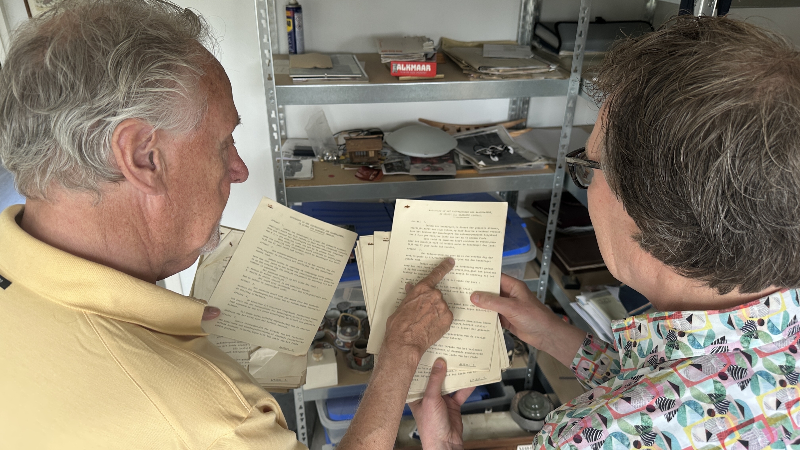 Twee mensen bekijken samen enkele getypte documenten in een ruimte met metalen planken gevuld met papieren en voorwerpen.