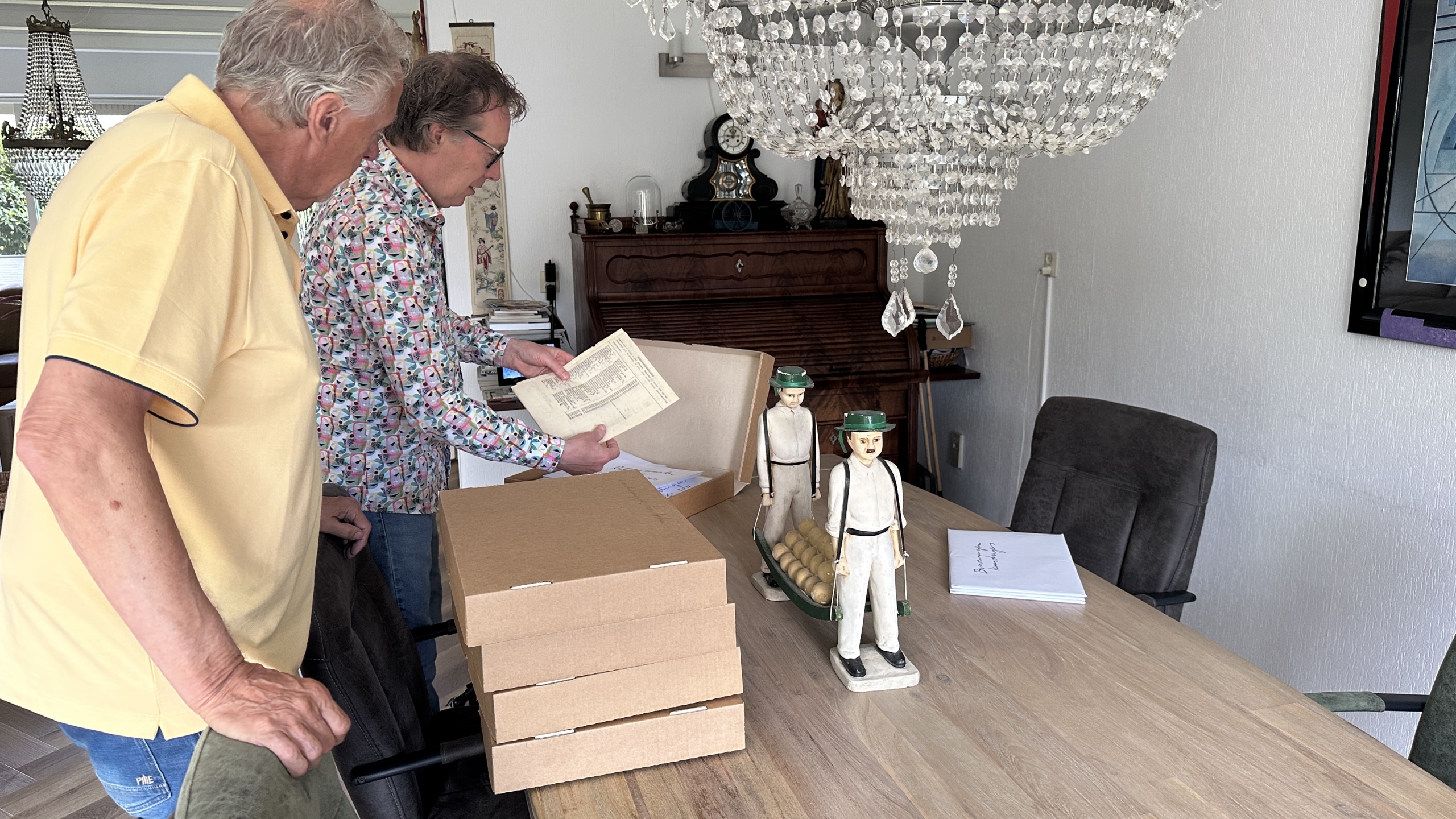 Twee mannen staan aan een tafel in een kamer gevuld met antieke meubels, waaronder een kristallen kroonluchter. Op de tafel liggen kartonnen dozen en papieren, terwijl een man een document bekijkt. Er staan ook twee vintage houten figuren op de tafel.