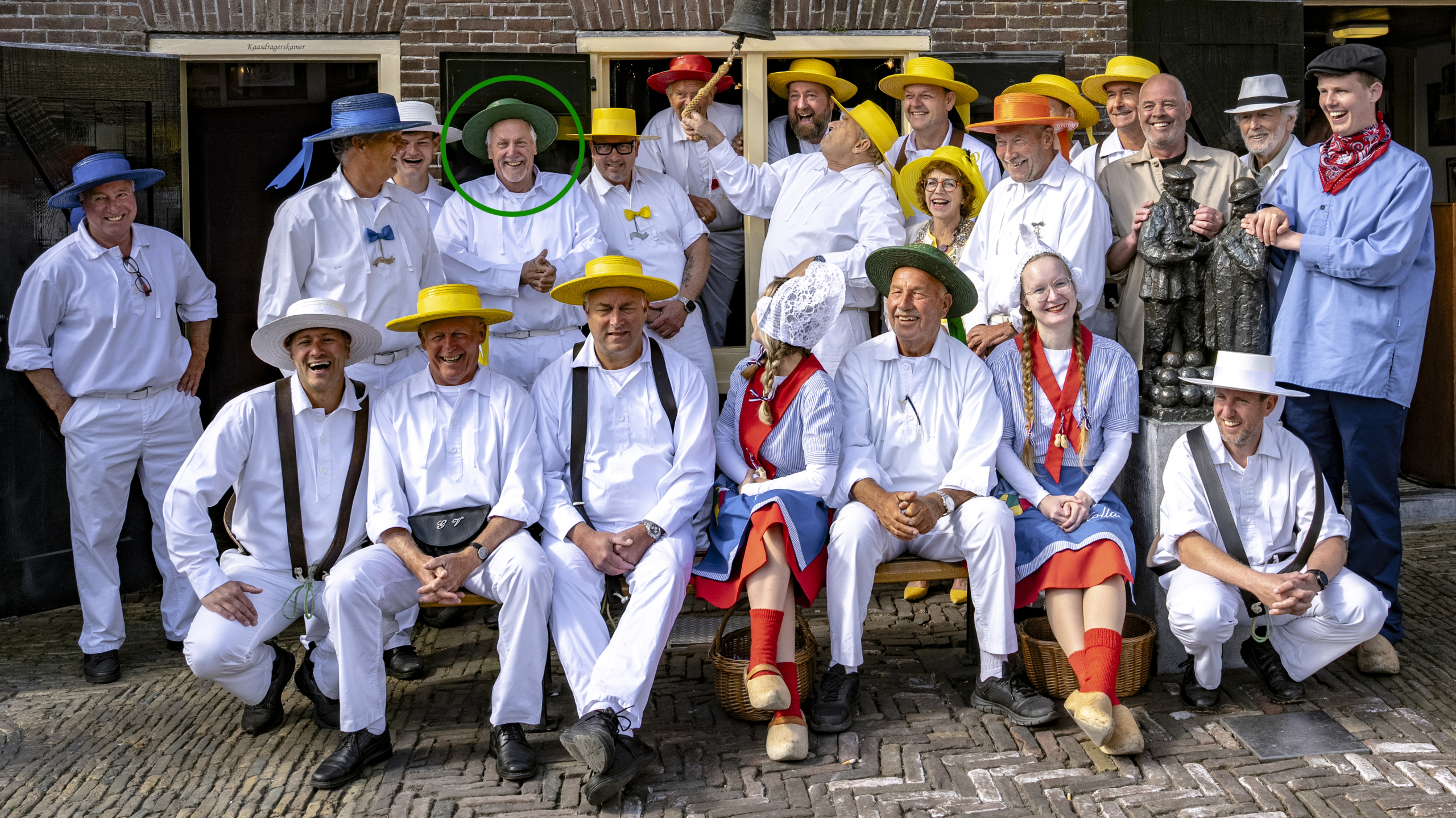 Een groep mensen in traditionele kledij staat en zit voor een gebouw. Ze dragen voornamelijk witte kleding met felgekleurde hoeden. Er staat een man in het midden met een groene cirkel om zijn hoofd, lachend naar de camera. De sfeer is vrolijk en feestelijk.
