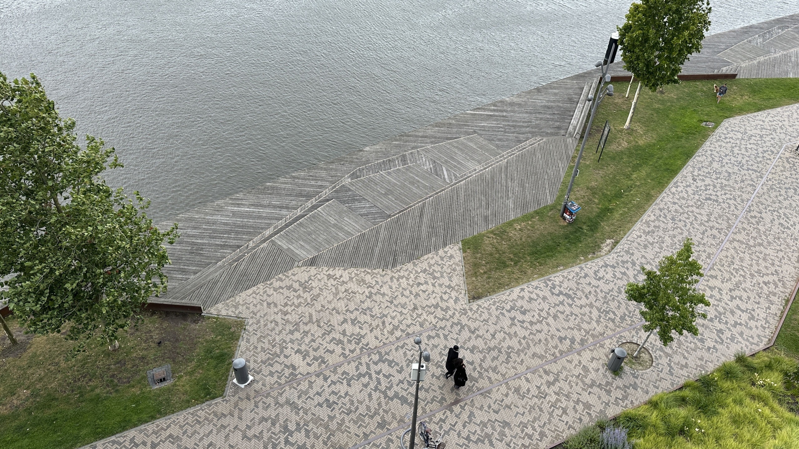 Een bovenaanzicht van een pad langs een waterkant met houten vlonders, grasvelden en bomen, waar enkele mensen wandelen.