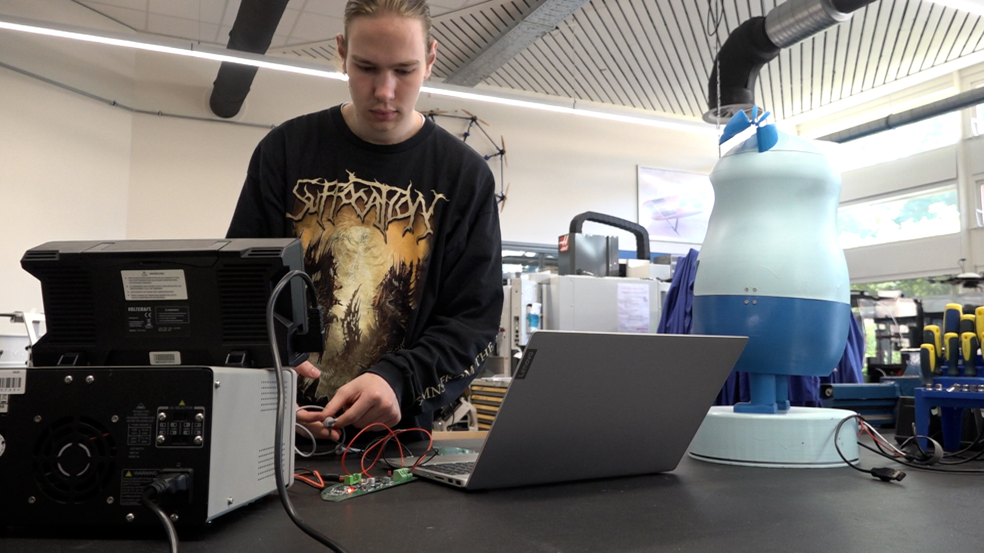 Man in a dark shirt works with electronic devices on a table, surrounded by technical equipment and a mannequin torso in a workshop setting.