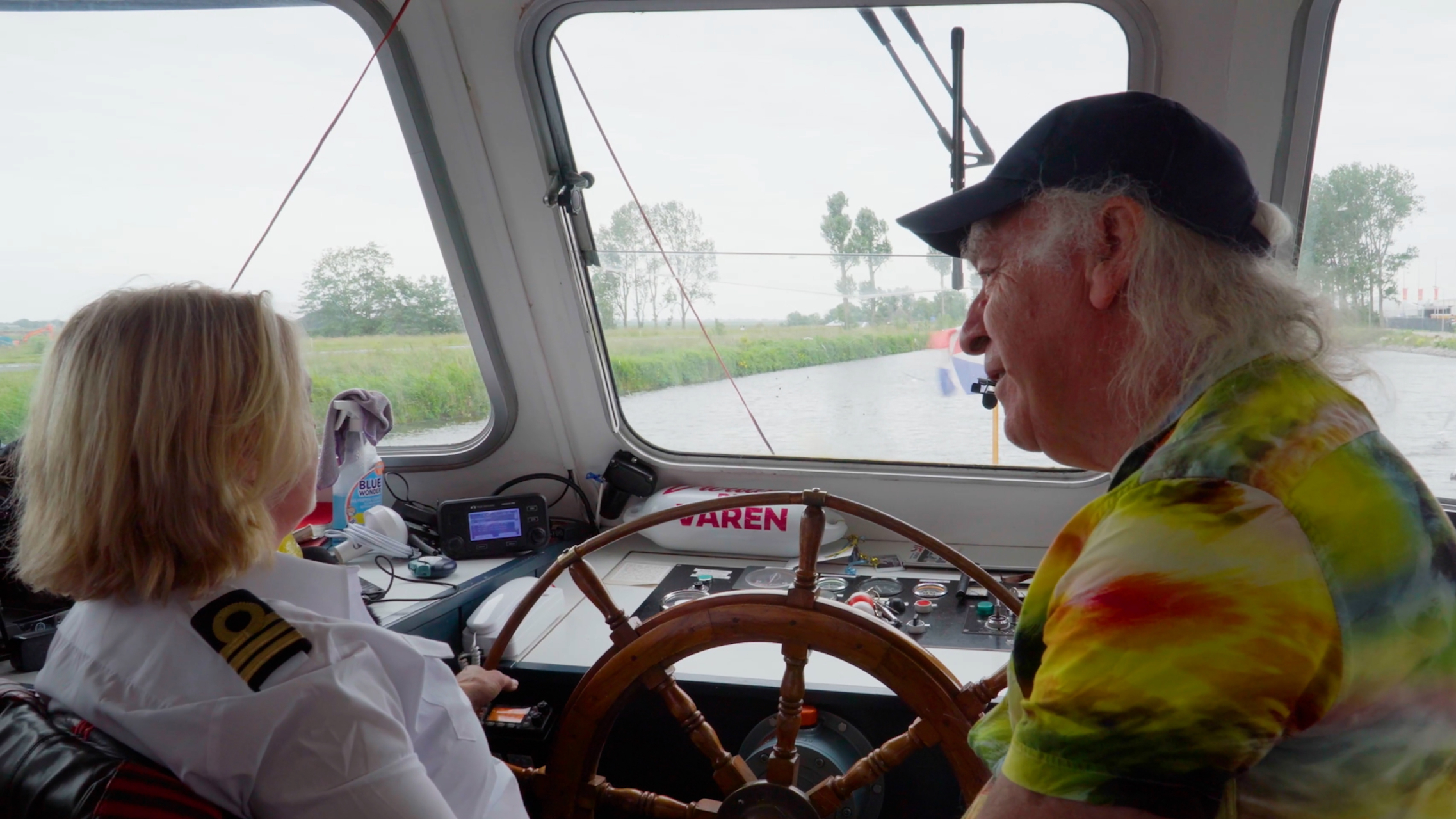 Twee personen zitten in de stuurhut van een boot; de ene persoon draagt een wit uniform met epauletten en de ander heeft een kleurrijk T-shirt aan. Ze kijken beiden naar voren, over een kanaal met groen landschap buiten de ramen.