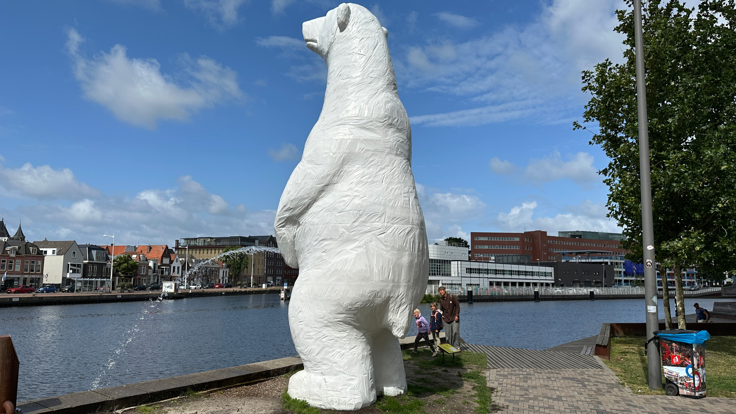 Een groot, witte ijsbeerstandbeeld staat aan de rand van een rivier met stedelijke gebouwen en een blauwe lucht op de achtergrond. Een vader en twee kinderen staan aan de voet van het beeld.