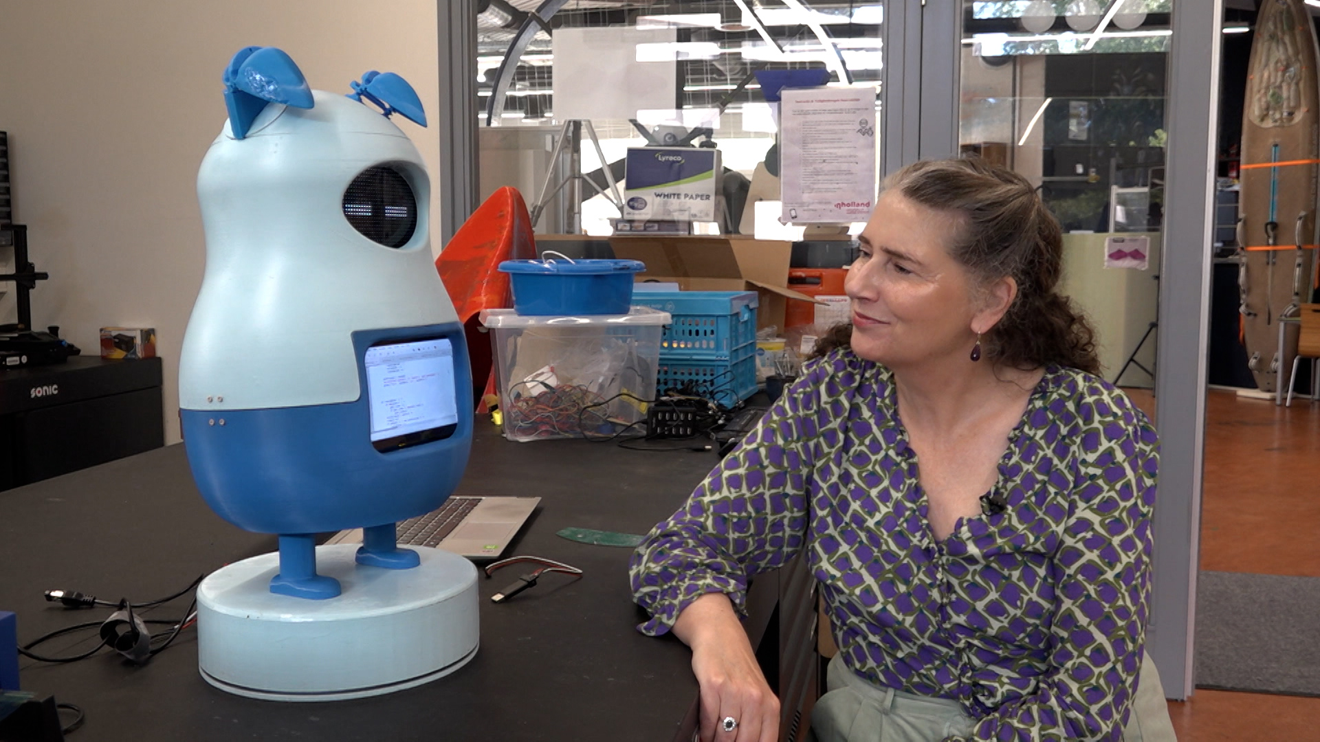 Een vrouw met lang haar kijkt naar een blauw robotachtig apparaat met oren, dat op een tafel in een werkplaats staat.