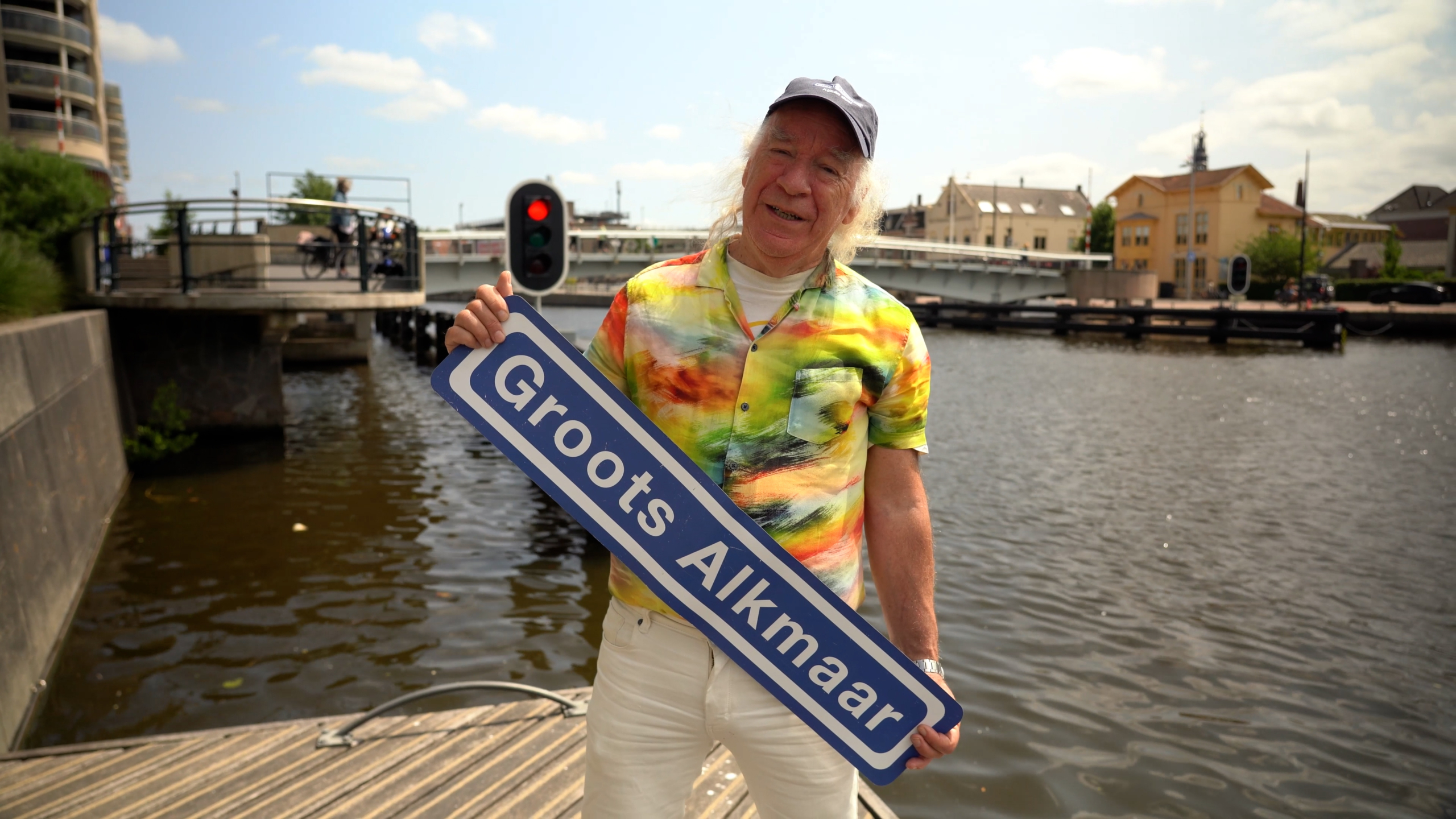 Een persoon met een kleurrijk overhemd staat aan het water en houdt een blauw bord met de tekst "Groots Alkmaar" vast.