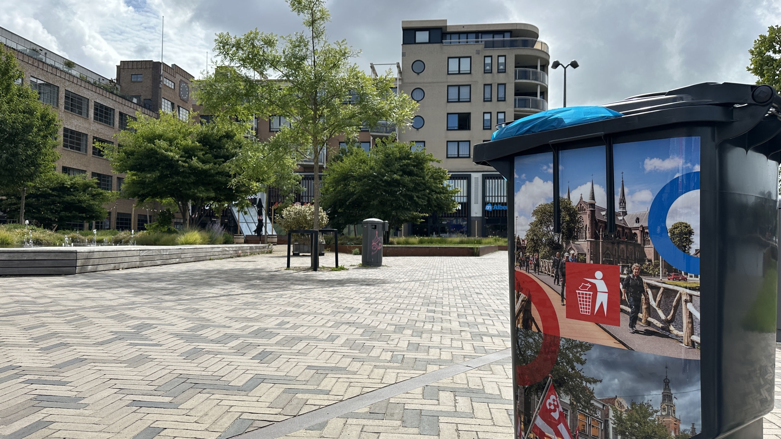 Stadsplein met moderne gebouwen op de achtergrond en een prullenbak op de voorgrond met afbeeldingen van een kerk en mensen die afval weggooien.