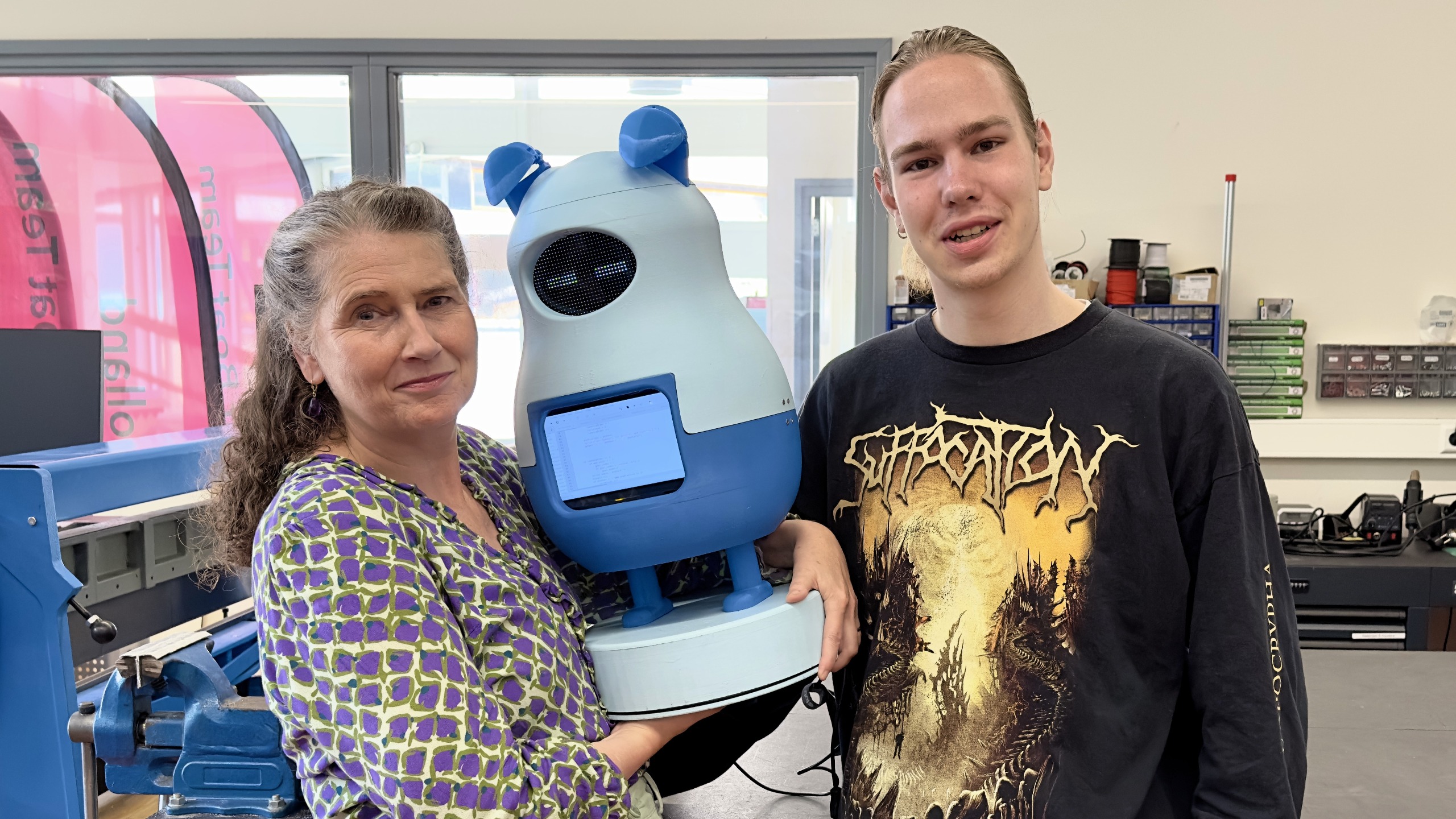 Een vrouw en een jongeman poseren met een blauw robotapparaat in een werkplaatsomgeving, met gereedschap en elektronische componenten op de achtergrond.
