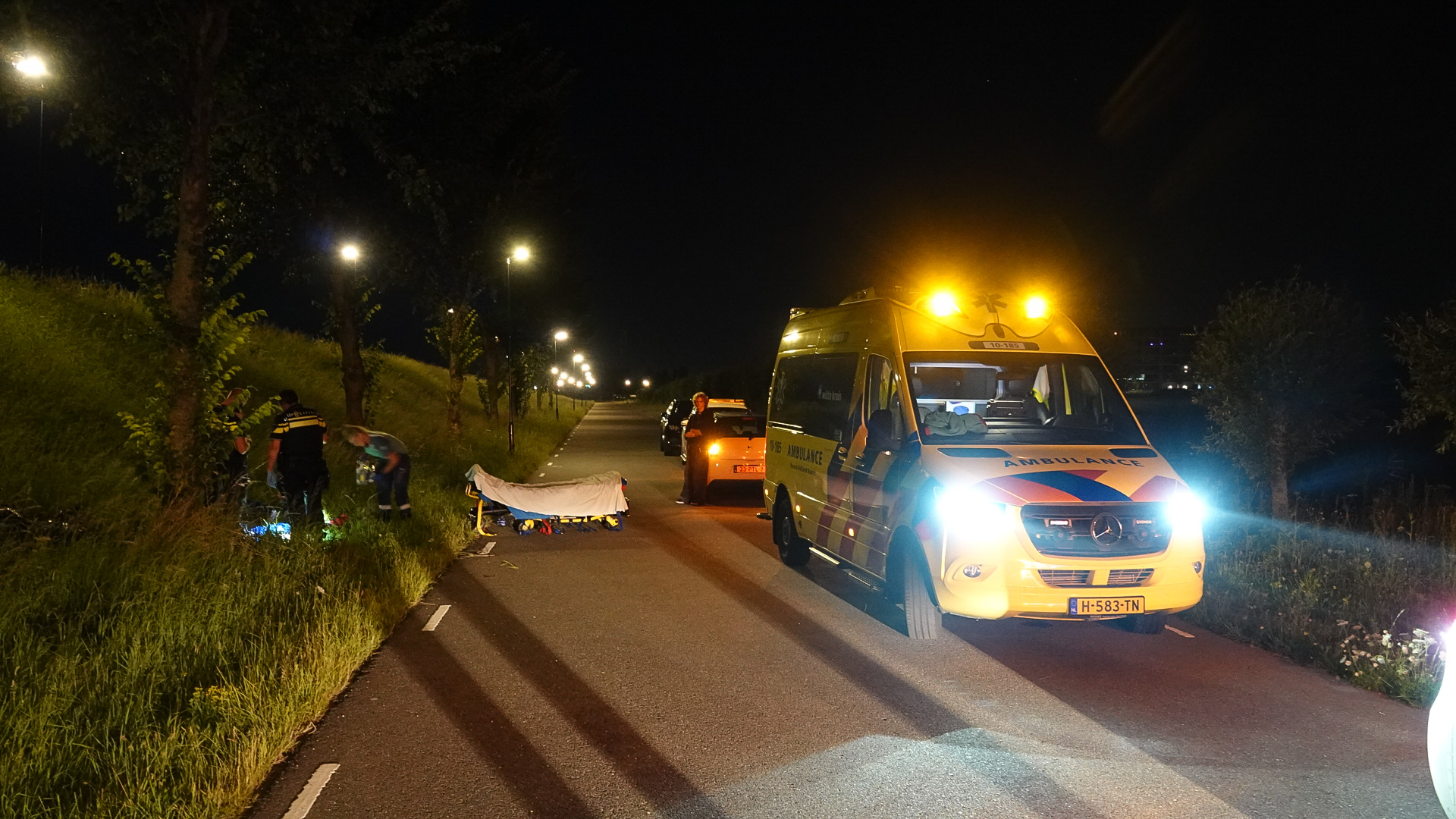 Een ambulance en politieagenten bij nacht op een weg, met noodverlichting aan en reddingsmateriaal op het asfalt uitgestald naast het voertuig.