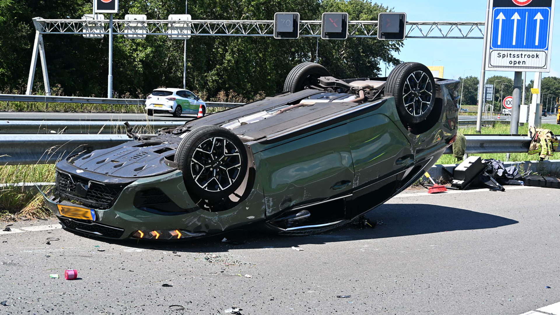 Een groene auto ligt ondersteboven op een snelweg naast een vangrail, met verkeersborden en een andere auto op de achtergrond.